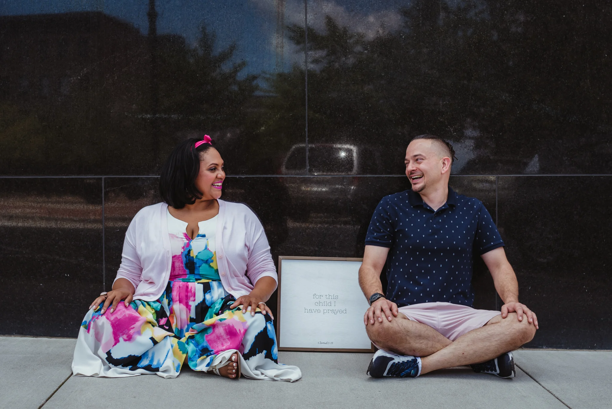 The parents to be laugh and sit by a sign that says “for this child I have prayed” outside in Durham during their baby announcement photo session with Rose Trail Images.