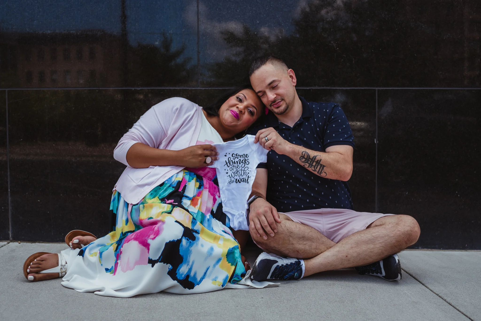 The parents to be sit on the sidewalk holding a onesie that says “some things are worth the wait” outside in Durham during their baby announcement photo session with Rose Trail Images.