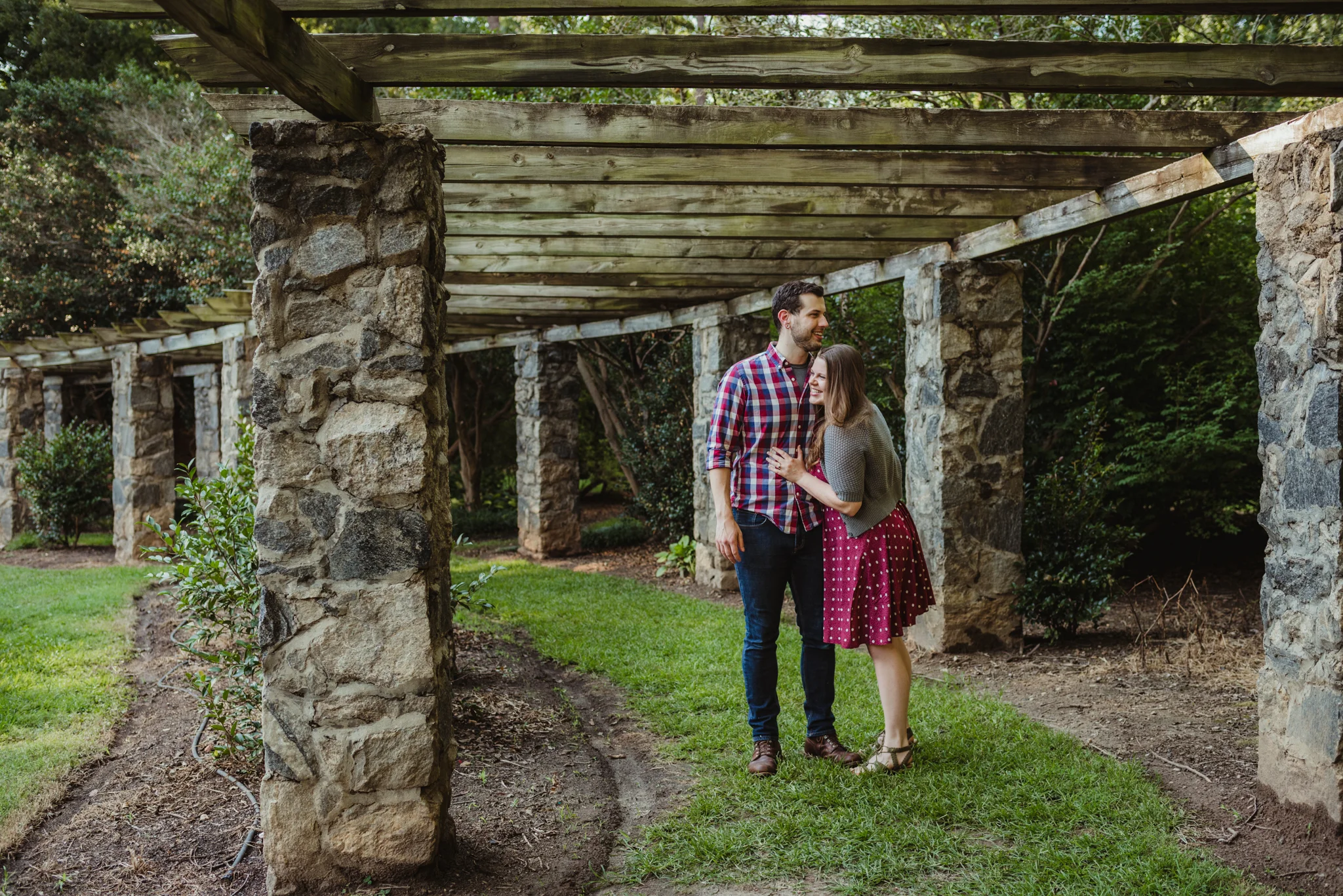 The bride and groom to be hold each other under the stone trellis at the Raleigh Rose Garden during their engagement session with Rose Trail Images.