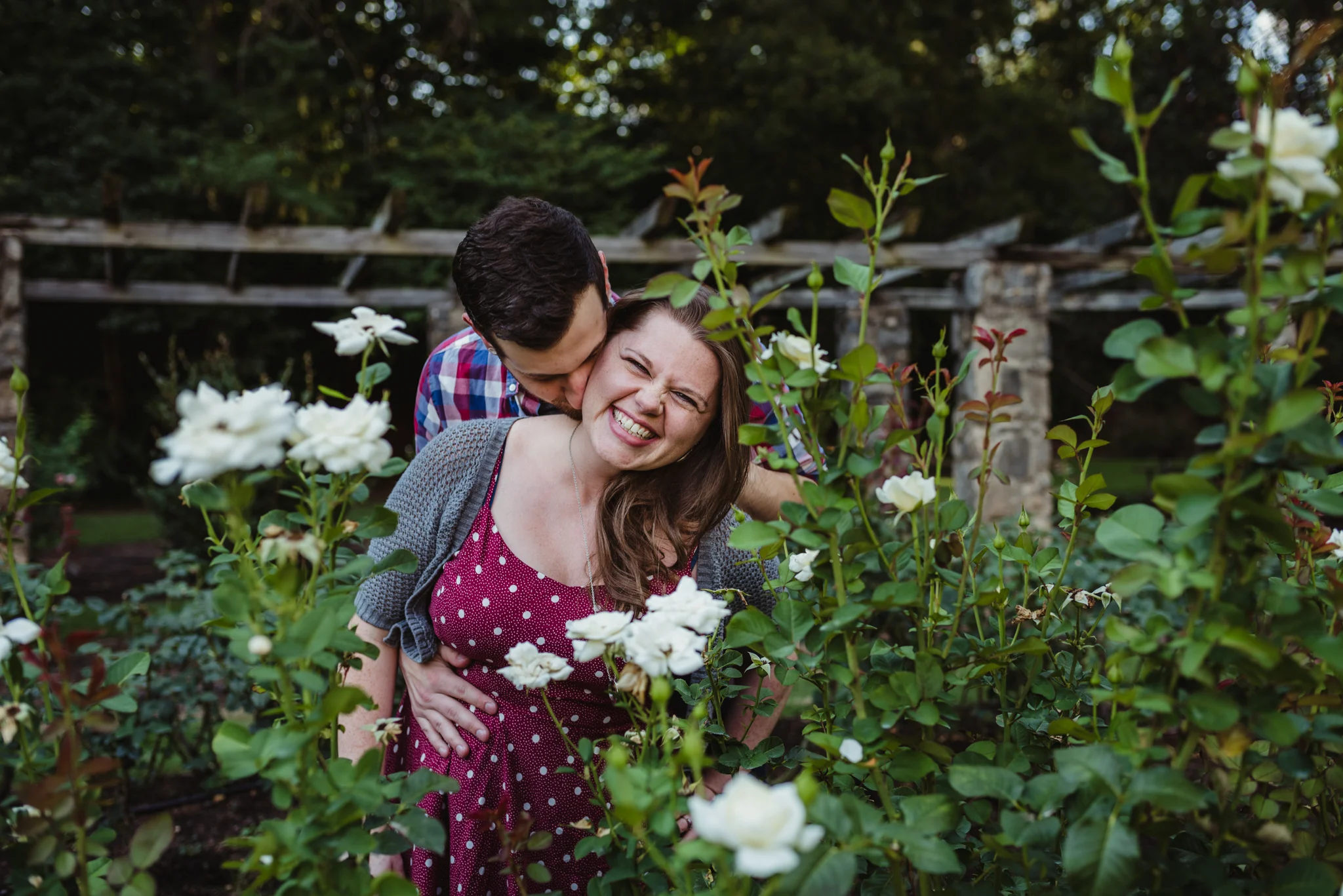 The groom kisses his bride to be on the neck while at the Raleigh Rose Garden during their engagement session with Rose Trail Images.