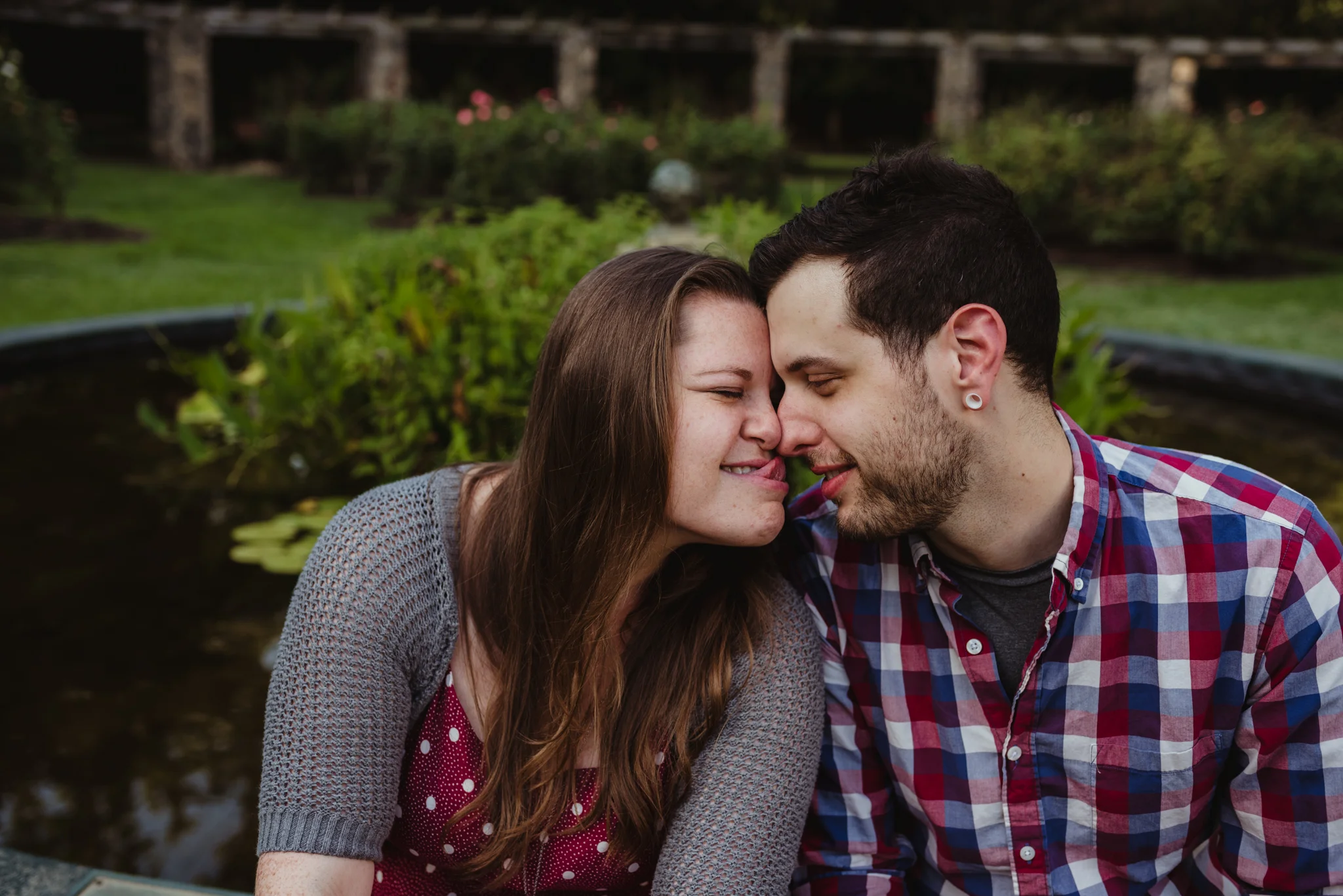 The bride and groom to be touch foreheads while she tries to lick his nose by the pond at the Raleigh Rose Garden during their engagement session with Rose Trail Images.