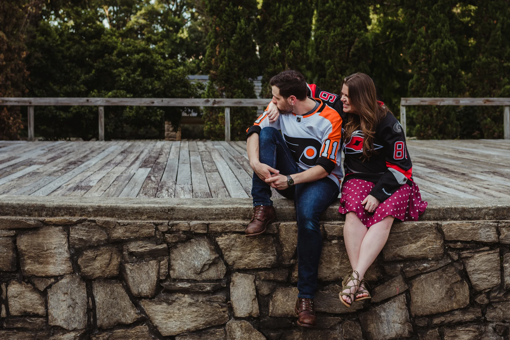 The bride and groom to be, in their Flyers and Hurricanes hockey jerseys, sit on the Little Theater stage at the Raleigh Rose Garden during their engagement session with Rose Trail Images.