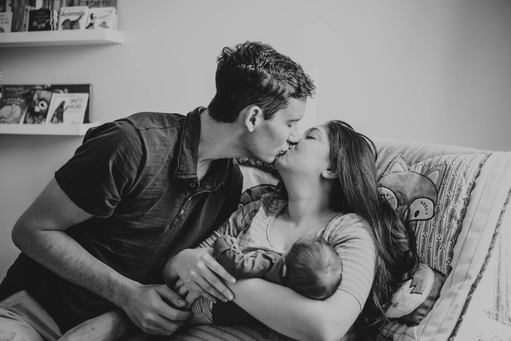 The new parents kiss in the chair in the nursery while holding their newborn in Wake Forest, a black and white image by Rose Trail Images.