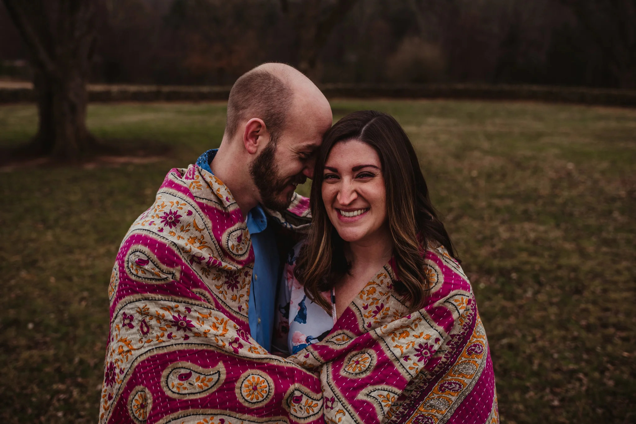 The parents to be snuggle under a pink blanket to represent their baby girl on the way, picture taken by Rose Trail Images at Joyner Park in Wake Forest, NC.
