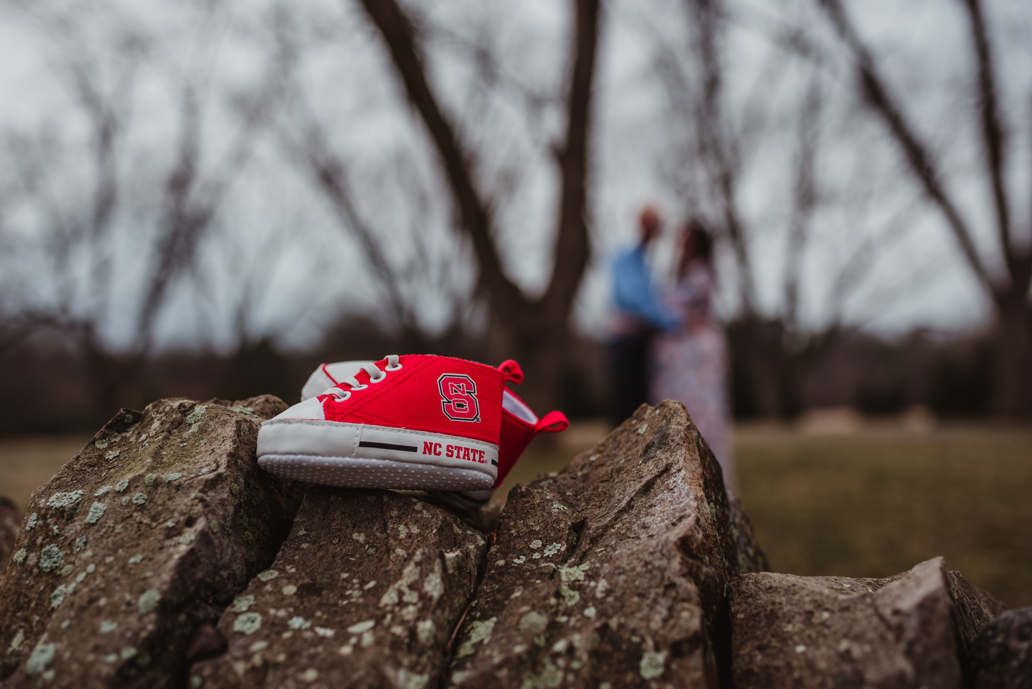 The parents to be show hug in the distance while they show off baby girl’s new NC State booties, picture taken by Rose Trail Images at Joyner Park in Wake Forest, NC.