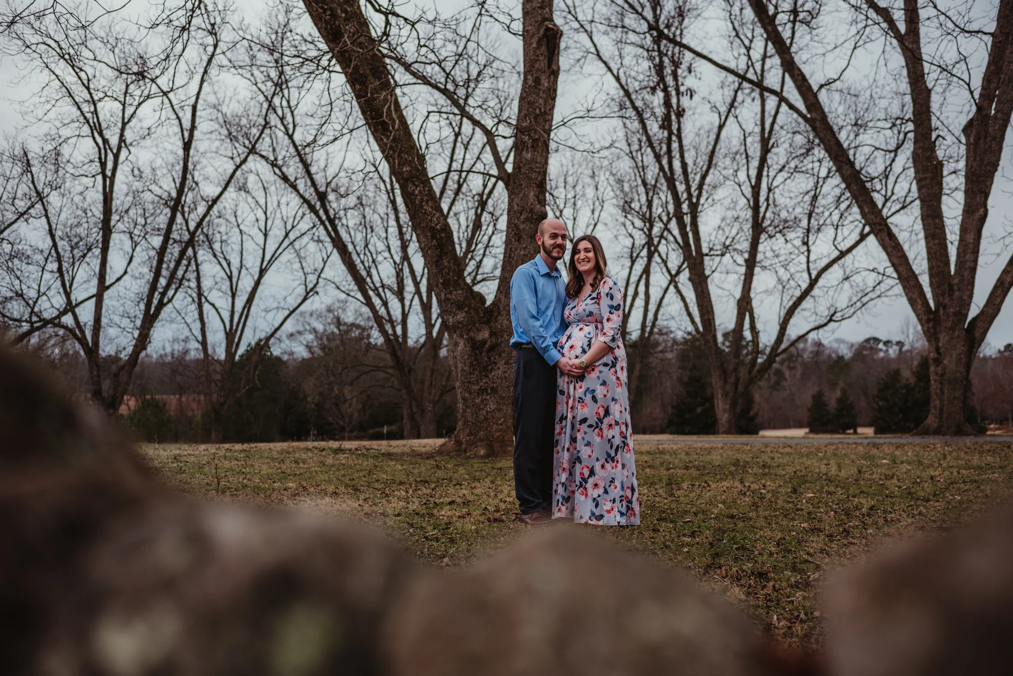 The parents to be hold each other and the baby bump in the pecan grove, picture taken by Rose Trail Images at Joyner Park in Wake Forest, NC.