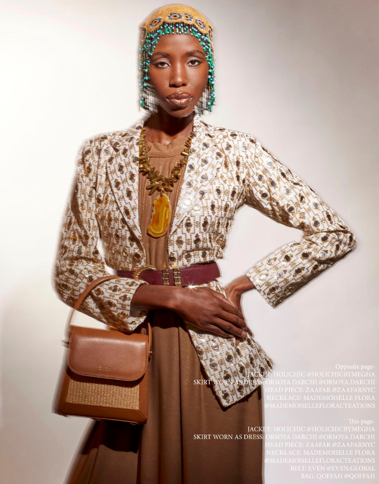 A woman wearing a beige patterned jacket, brown skirt, and accessories including a hat with beads, a large necklace with a yellow pendant, and a brown handbag, standing against a plain background.