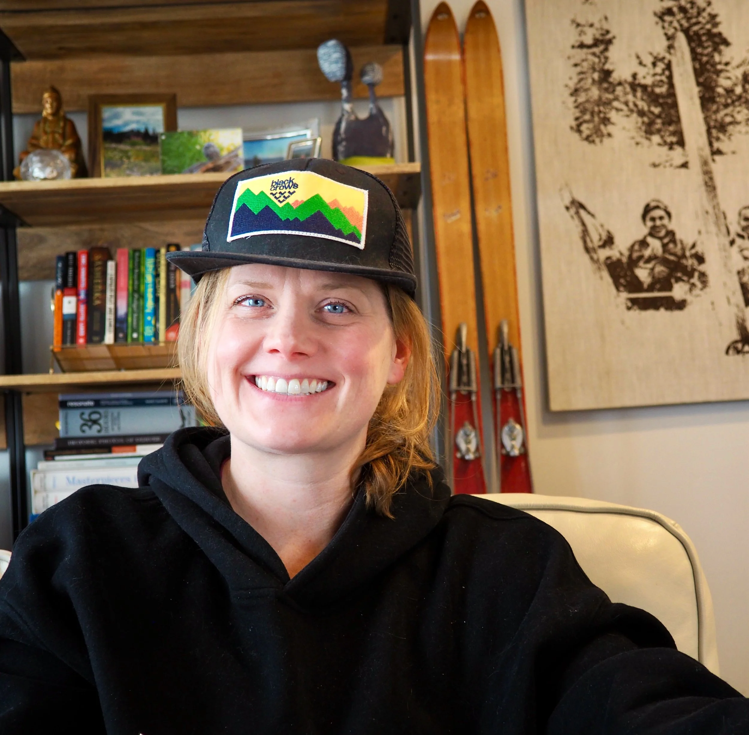 picture of kendra smiling in front of shelves wearing a black baseball hat and black sweatshirt