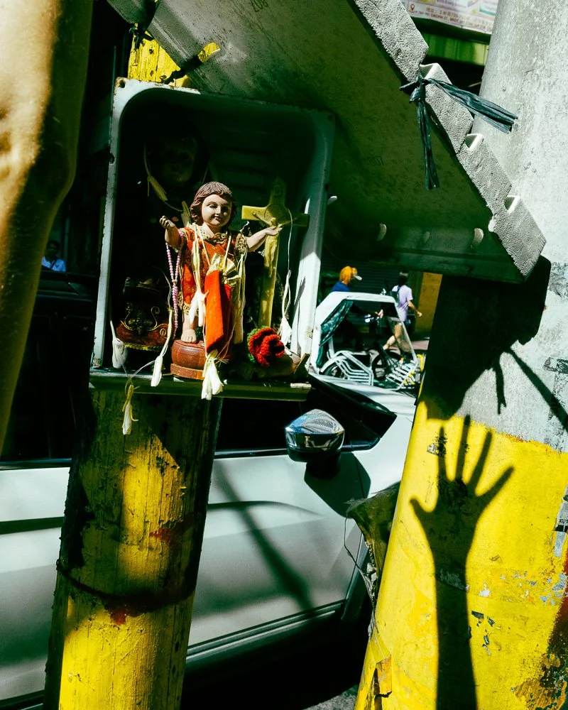 A religious statue in a roadside shrine on a yellow post, with the shadow of a hand on the wall