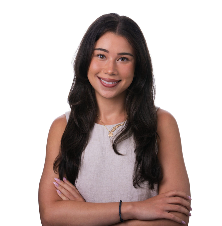 A young woman with long dark hair, wearing a sleeveless light gray top, smiling with arms crossed against a white background.