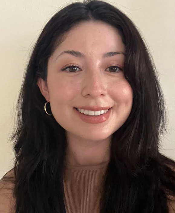 A young woman with long, dark hair smiling at the camera, wearing a brown top and hoop earrings against a plain beige background.