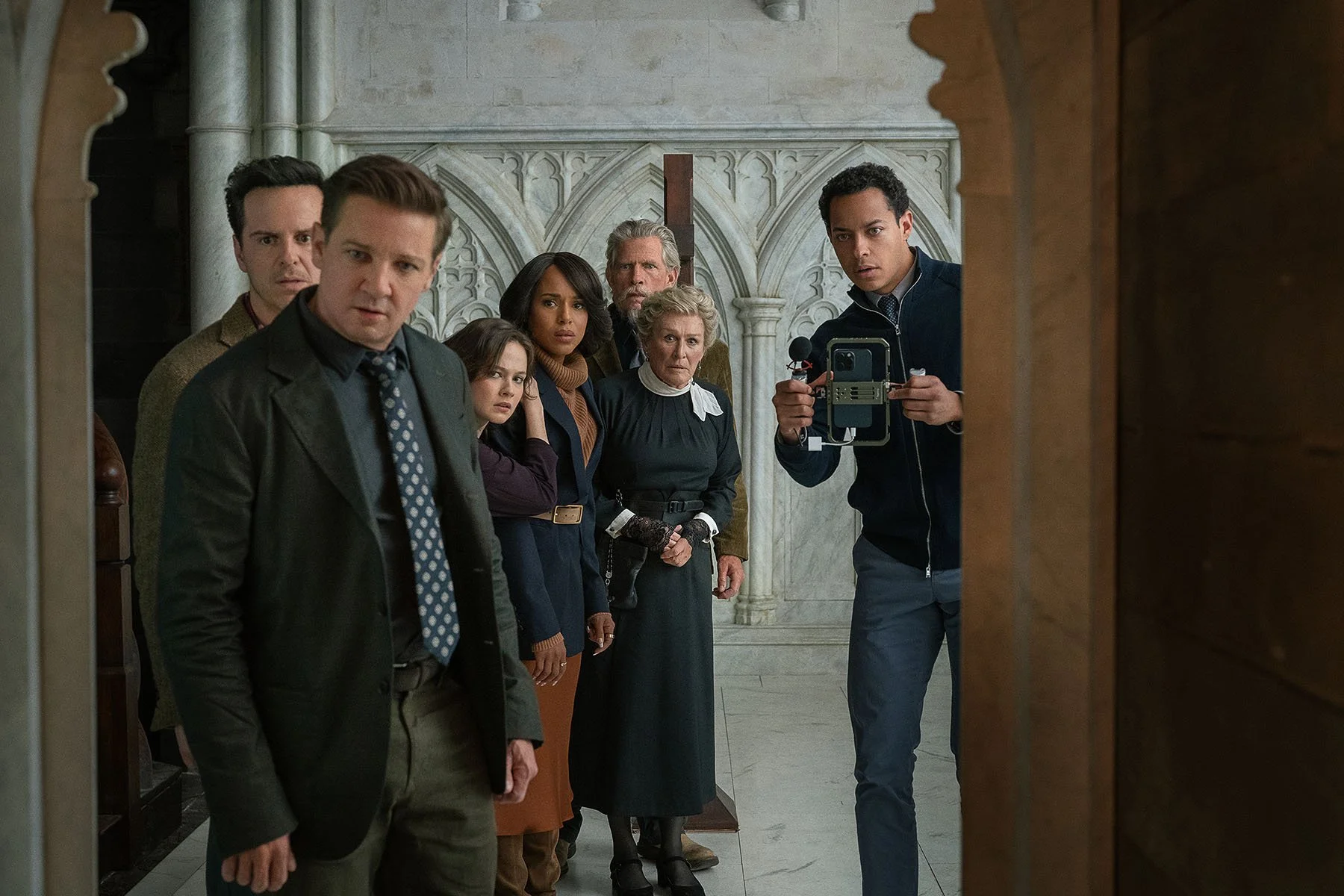 Seven people stare into an entryway, with confused looks. The man on the far right holds a camera.