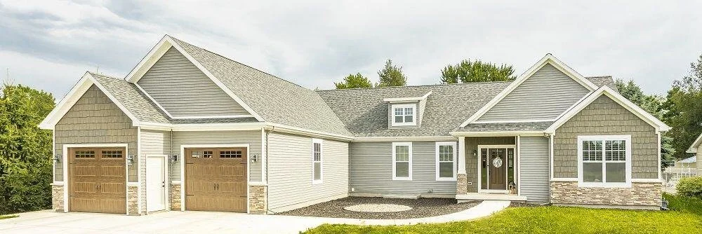 A large modern suburban house with gray siding, a gabled roof, and a two-car garage, surrounded by a lawn and trees.