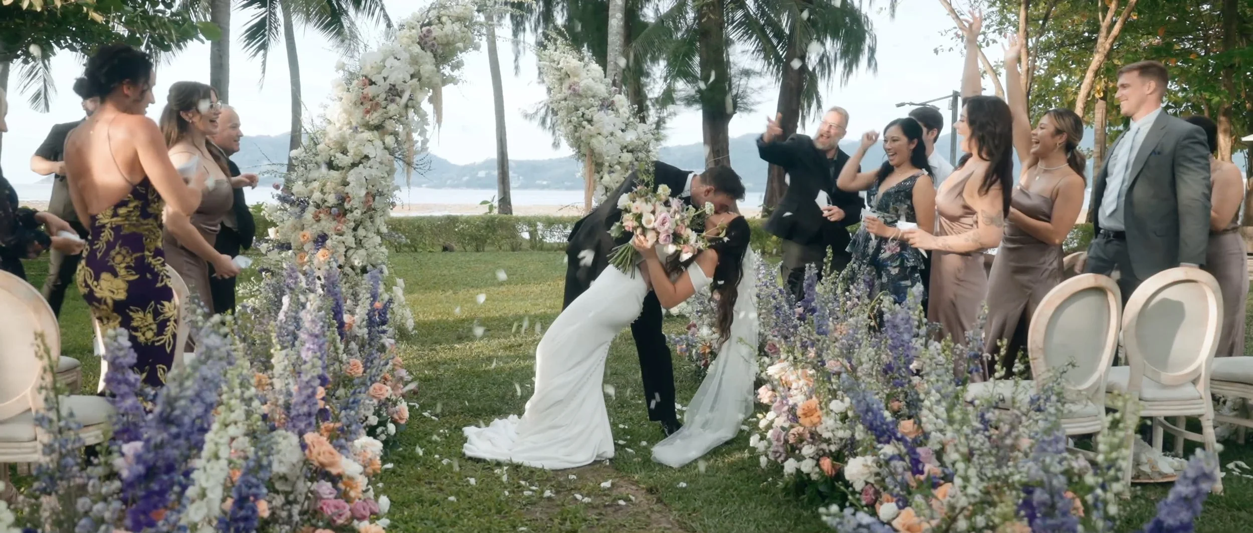 The couple at the Rosewood Phuket resort, sharing a dip kiss during the ceremony processional, as flower petals are tossed around them.