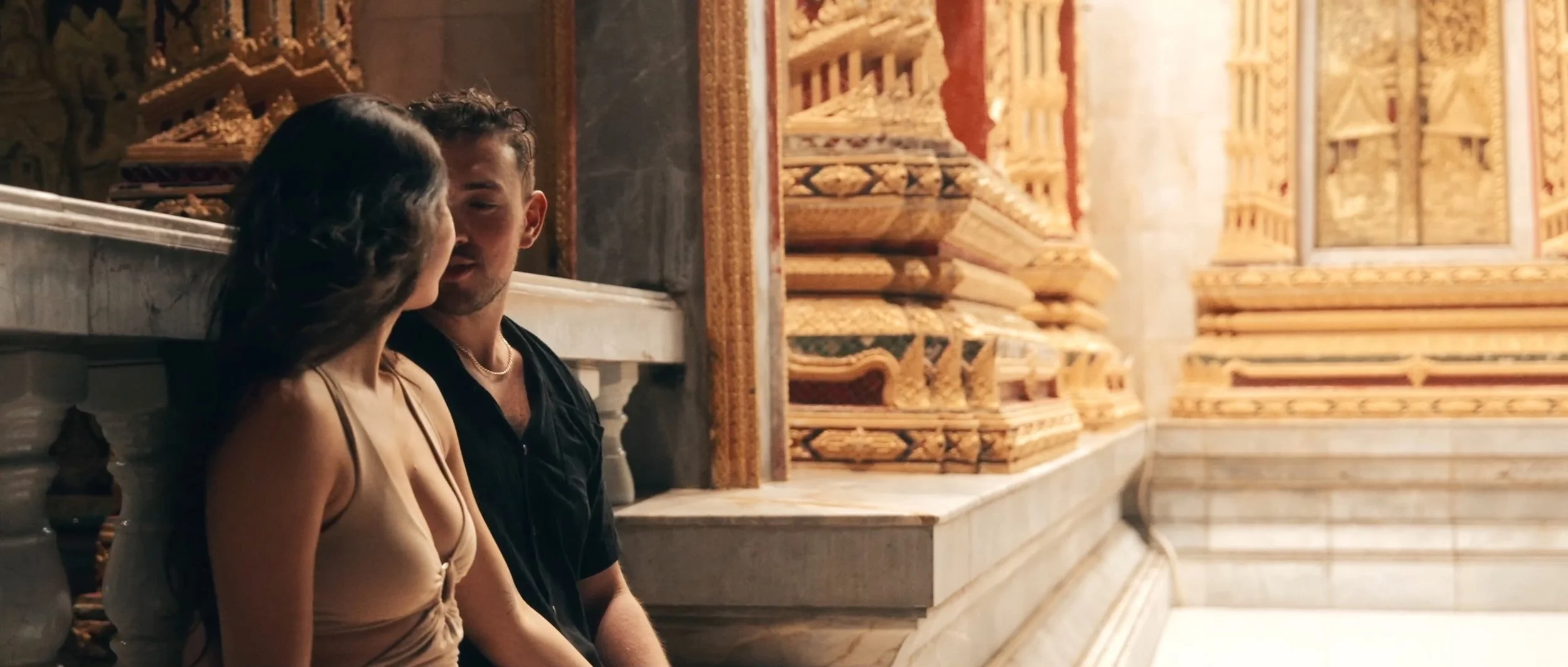 A couple sitting together on a bench at a temple in Thailand, surrounded by intricate architecture, in a quiet moment.