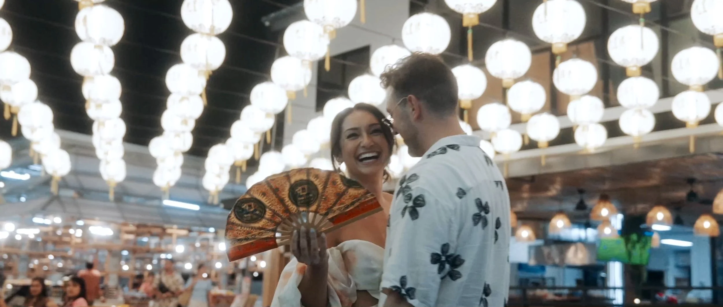 The couple standing together under glowing paper lanterns in a vibrant night market scene.