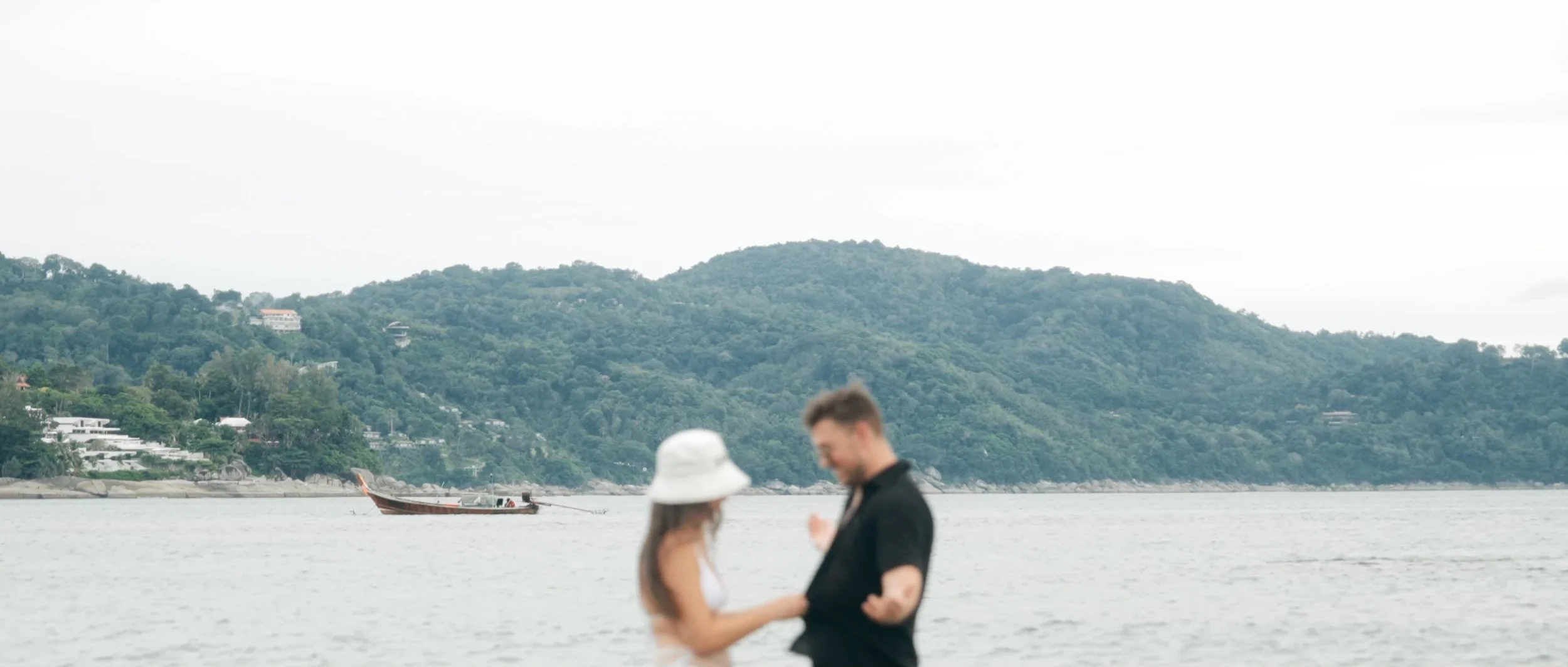A couple standing by the beach with lush greenery and a distant boat, capturing a serene coastal moment in Thailand