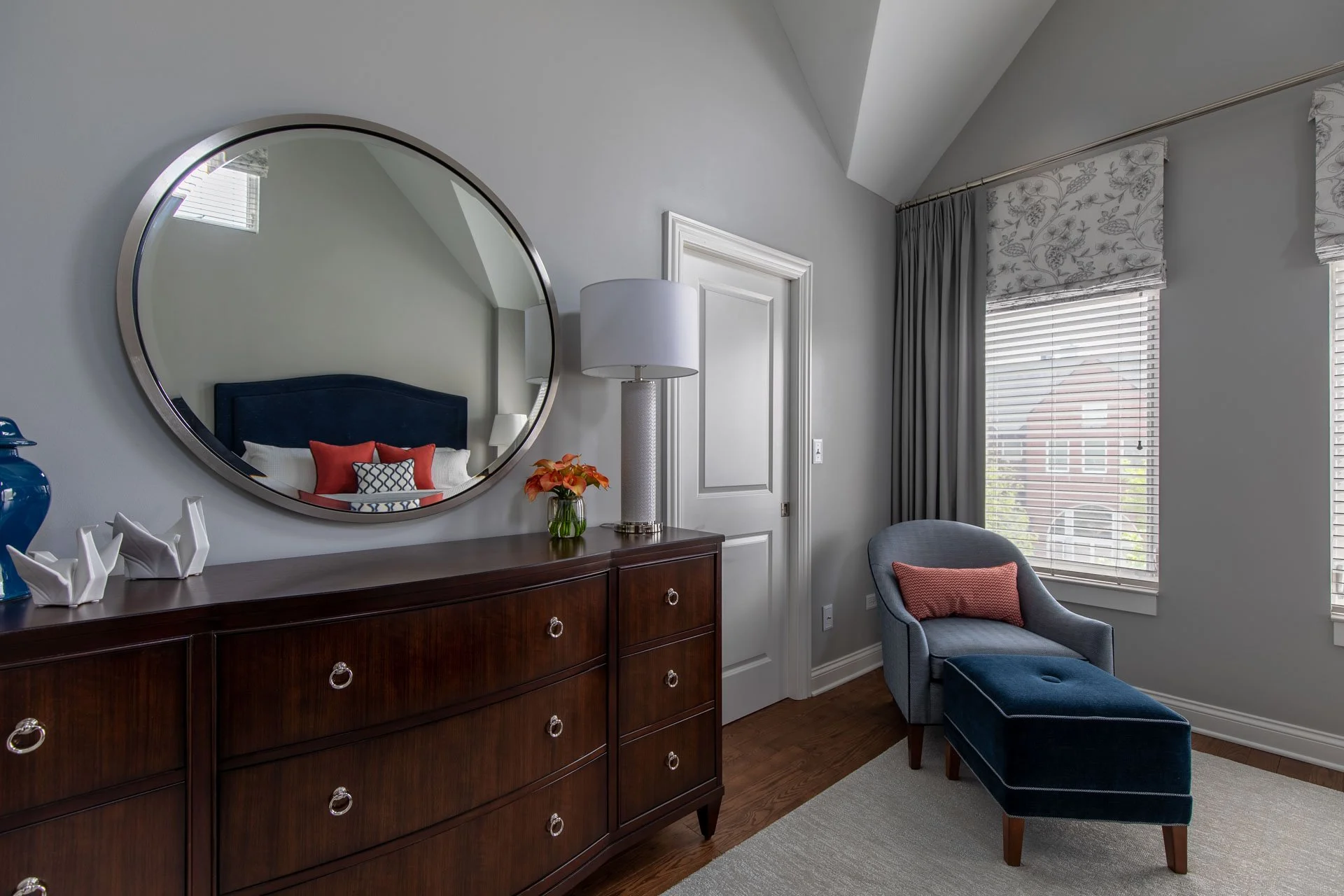 Primary bedroom in Des Plaines IL featuring a dark wood dresser with round mirror, modern table lamp, and upholstered seating with ottoman and orange accent pillow designed by Wendy Nobriga Interiors.