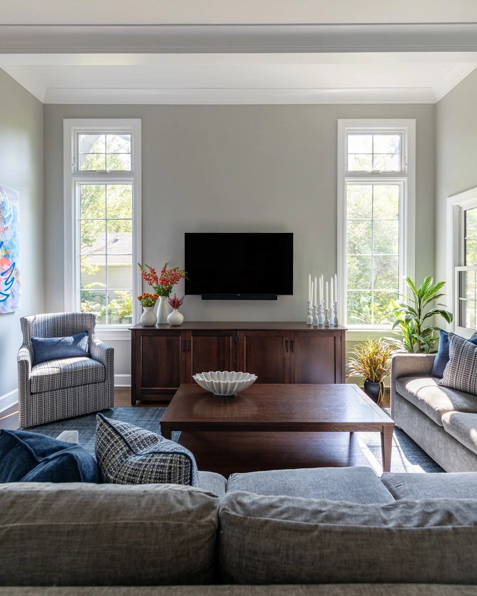 Family room in La Grange, IL with TV wall, wood console, flanking windows, accent chair, plants, and gray sectional, designed by Wendy Nobriga Interiors.