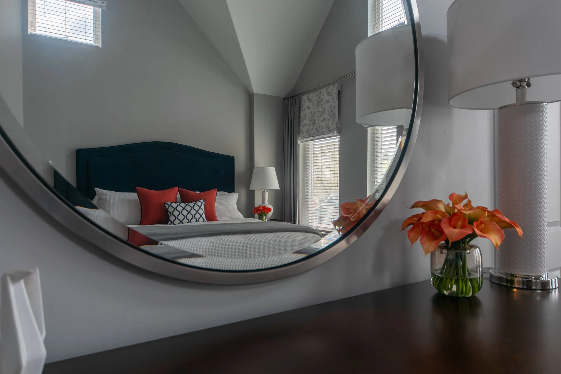 Primary bedroom in Des Plaines IL with a mirror reflecting the bed featuring a navy headboard, white bedding, and orange accent pillows, styled with a lamp and fresh orange flowers on a dark wood dresser, designed by Wendy Nobriga Interiors.