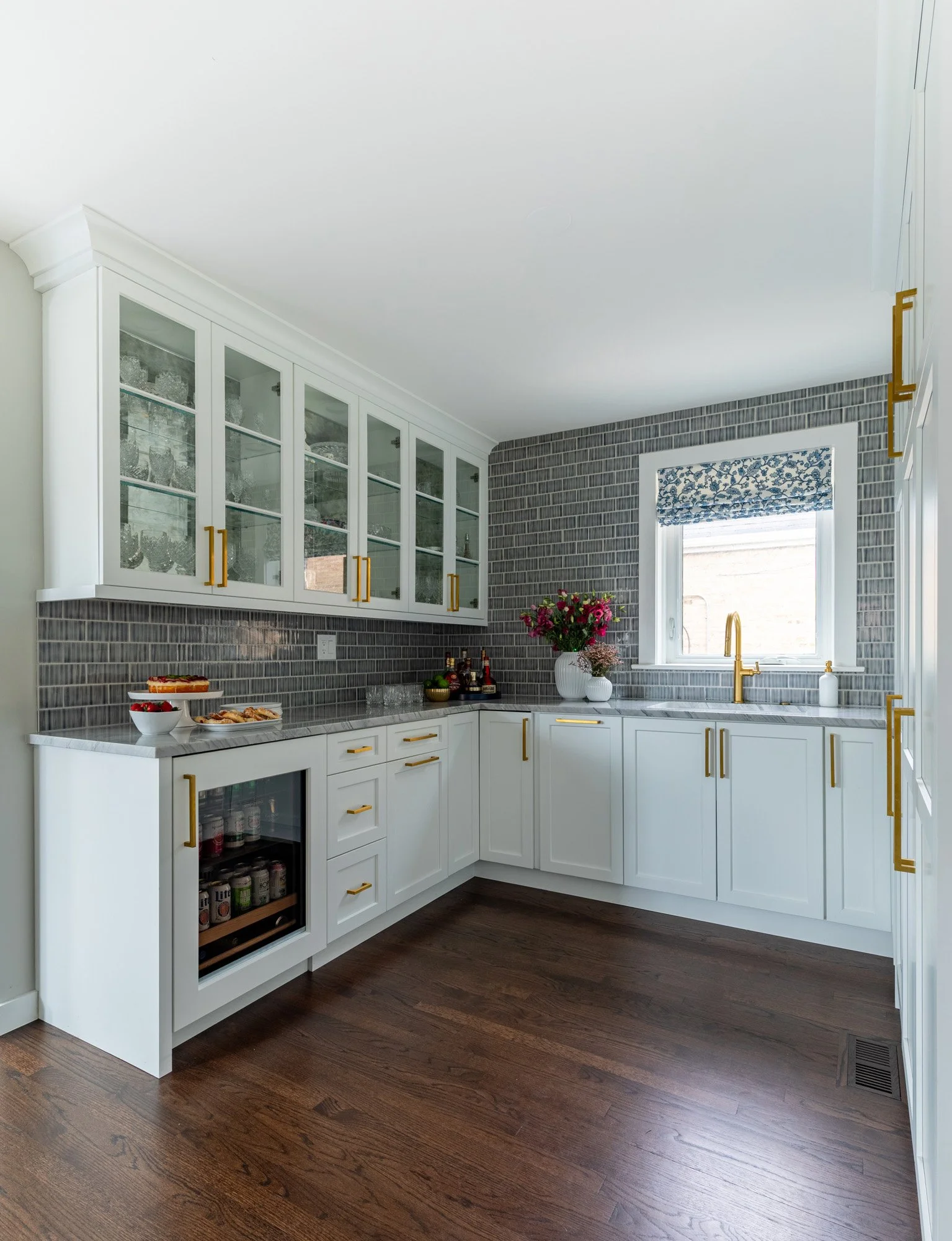 Custom beverage center in a North Edgebrook Chicago home featuring glass display cabinets, a beverage refrigerator, secondary sink, quartzite countertops, blue handcrafted tile backsplash, and custom cabinetry, designed by Wendy Nobriga Interiors.