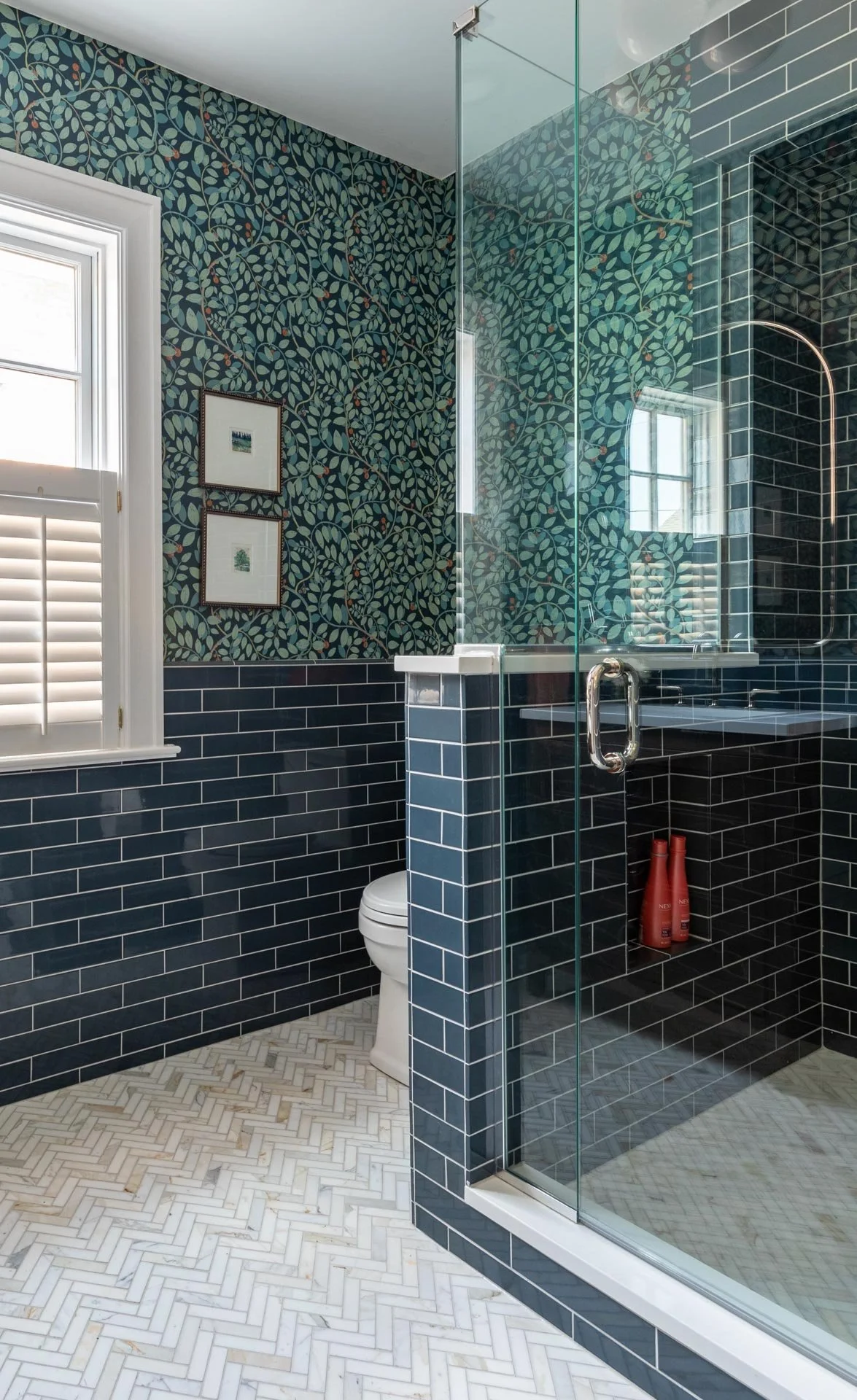 Lake Forest IL bathroom featuring glass shower enclosure, deep blue subway tile walls, botanical wallpaper, and herringbone marble floor, designed by Wendy Nobriga Interiors.