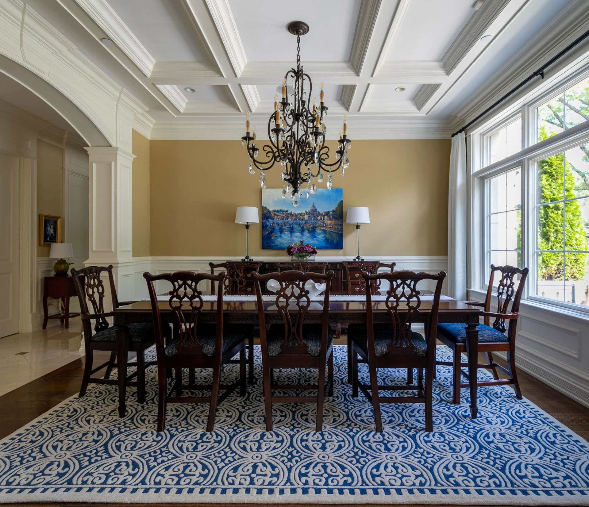 Lincolnwood IL dining room with coffered ceiling, wrought iron chandelier, large wood dining table, blue patterned rug, and artwork above the sideboard, designed by Wendy Nobriga Interiors.