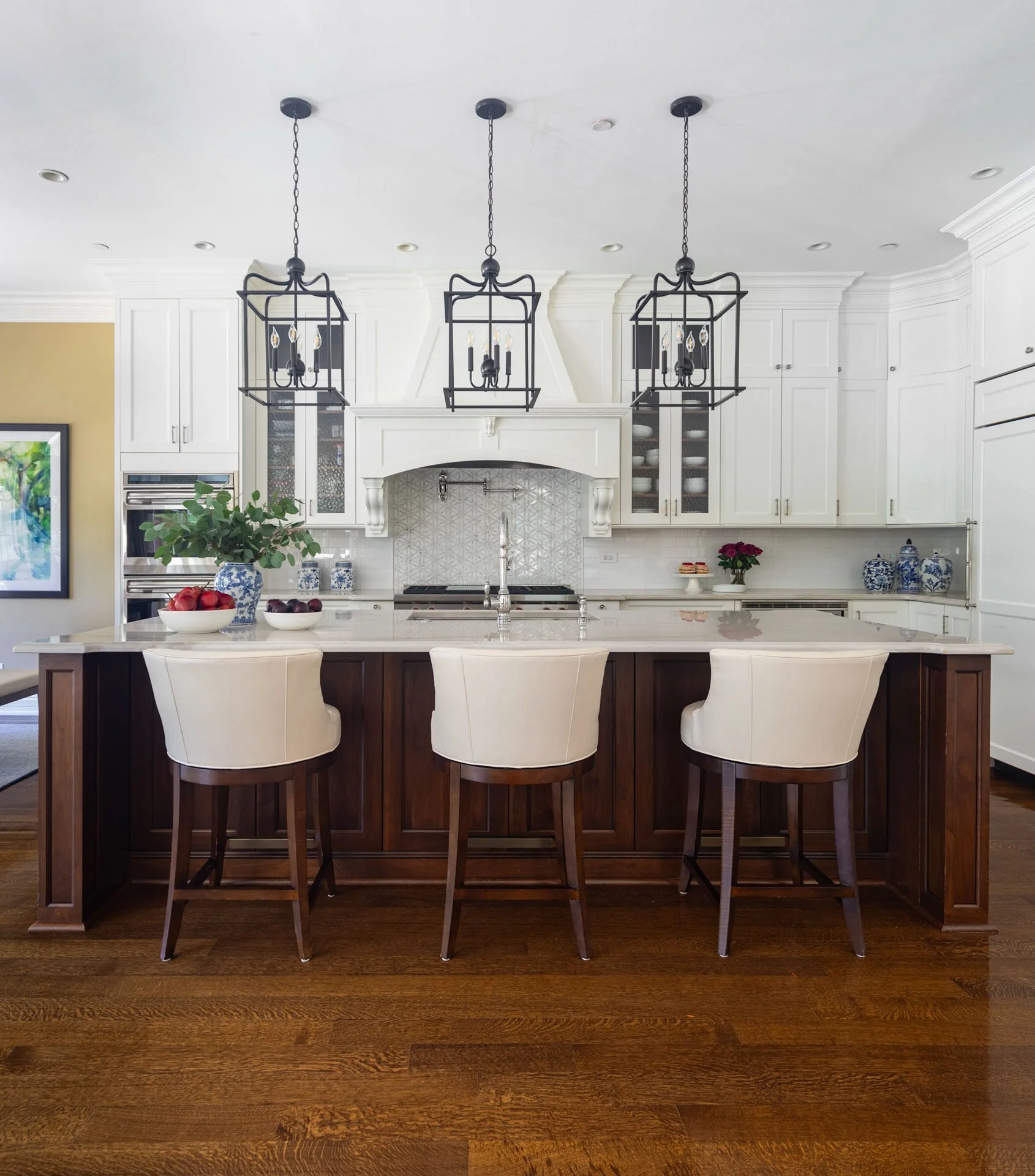 Lincolnwood IL kitchen with white cabinetry, dark wood island, quartzite countertops, and three black lantern pendant lights above white leather barstools, designed by Wendy Nobriga Interiors.