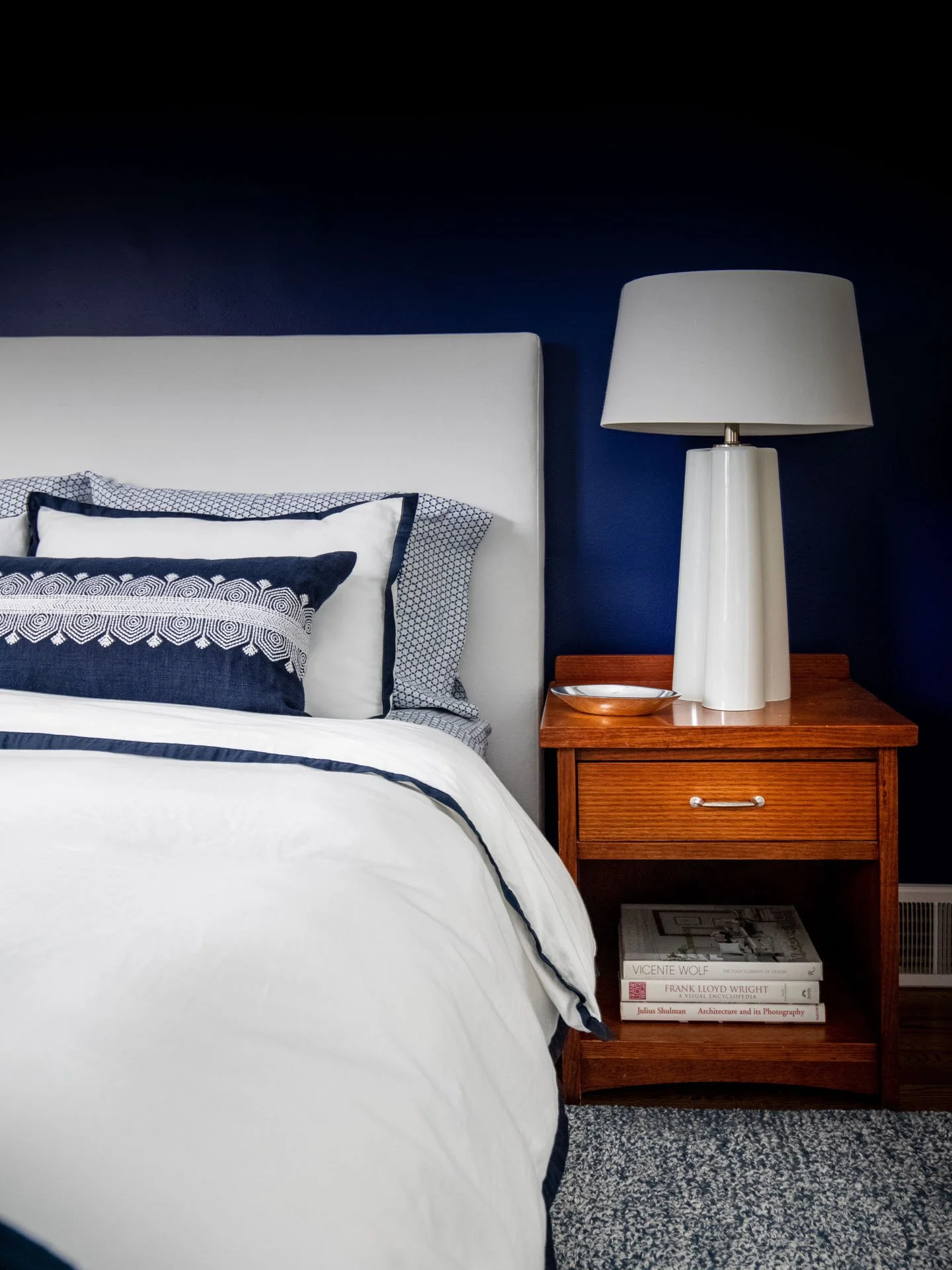 Primary bedroom in Edgebrook, Chicago, IL with navy accent wall, upholstered headboard, layered navy and white bedding, and wood nightstand with modern white lamp, designed by Wendy Nobriga Interiors.