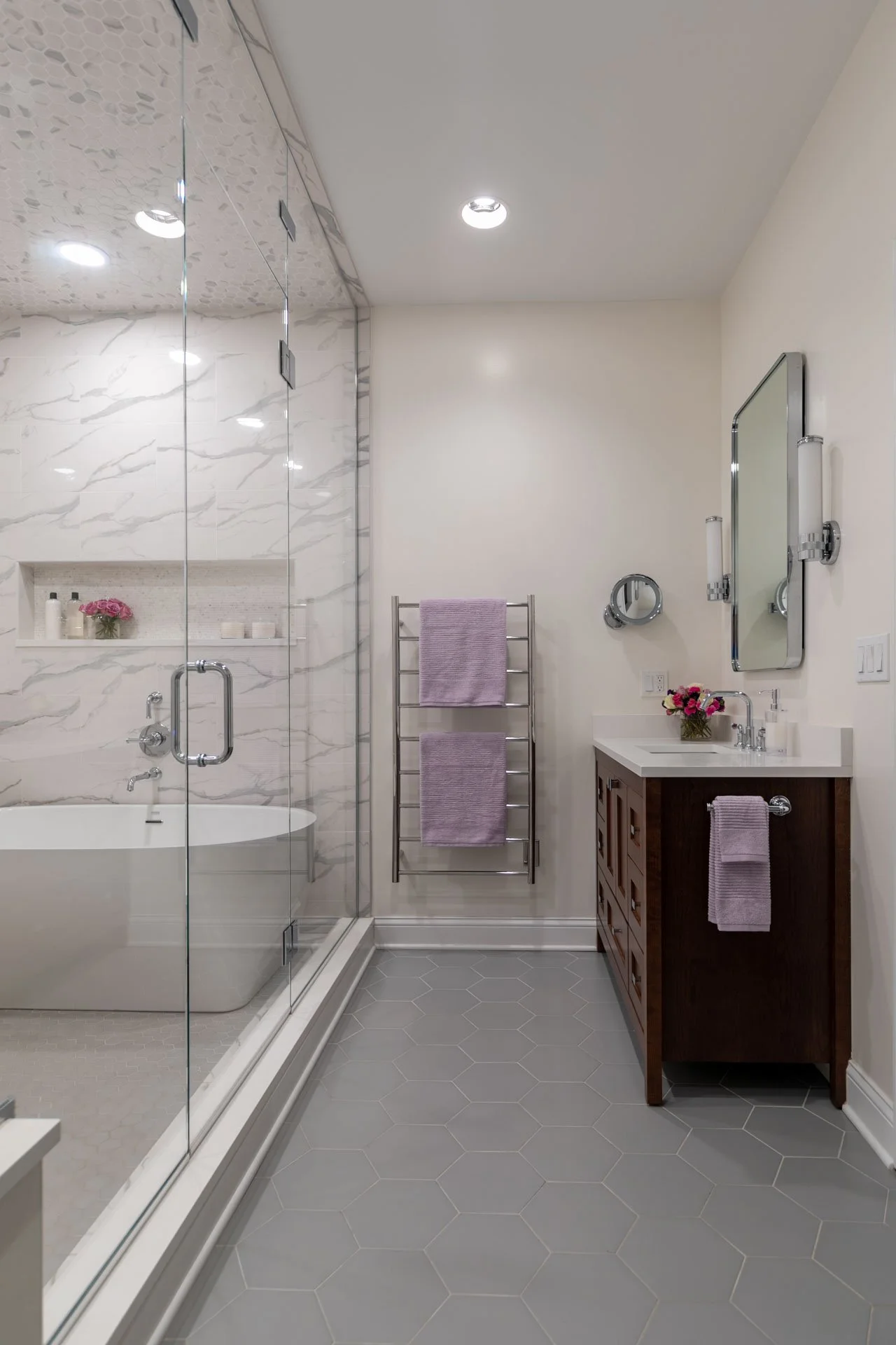 Lake View Chicago primary bathroom featuring a wood vanity with white countertop, polished chrome fixtures, lavender towels on a heated towel rack, and view into wet room with soaking tub and porcelain marble-look tile, designed by Wendy Nobriga Inte