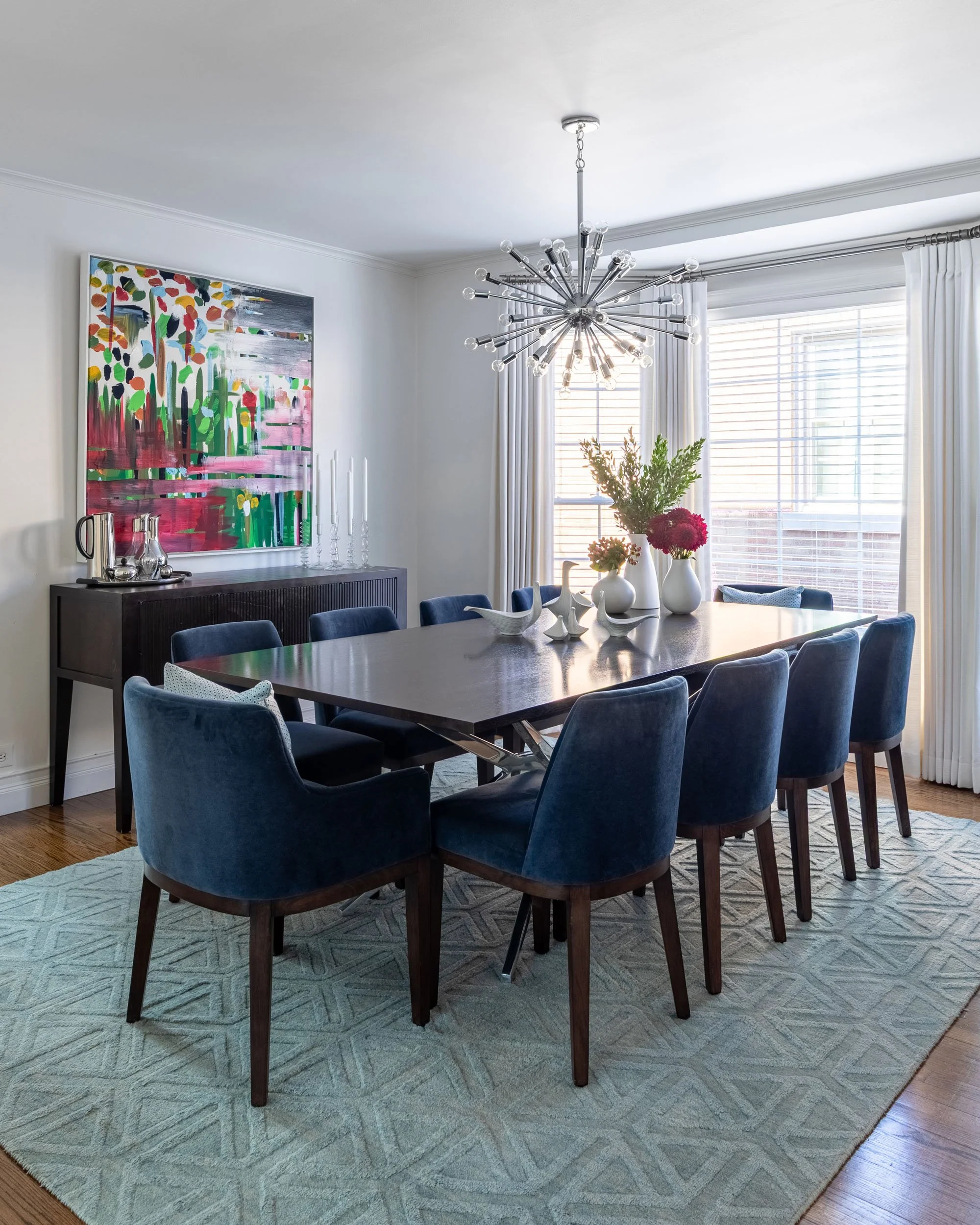 Dining room in Edgebrook, Chicago, IL with a modern dark wood table, blue velvet chairs, statement chandelier, and abstract artwork. Design by Wendy Nobriga Interiors.
