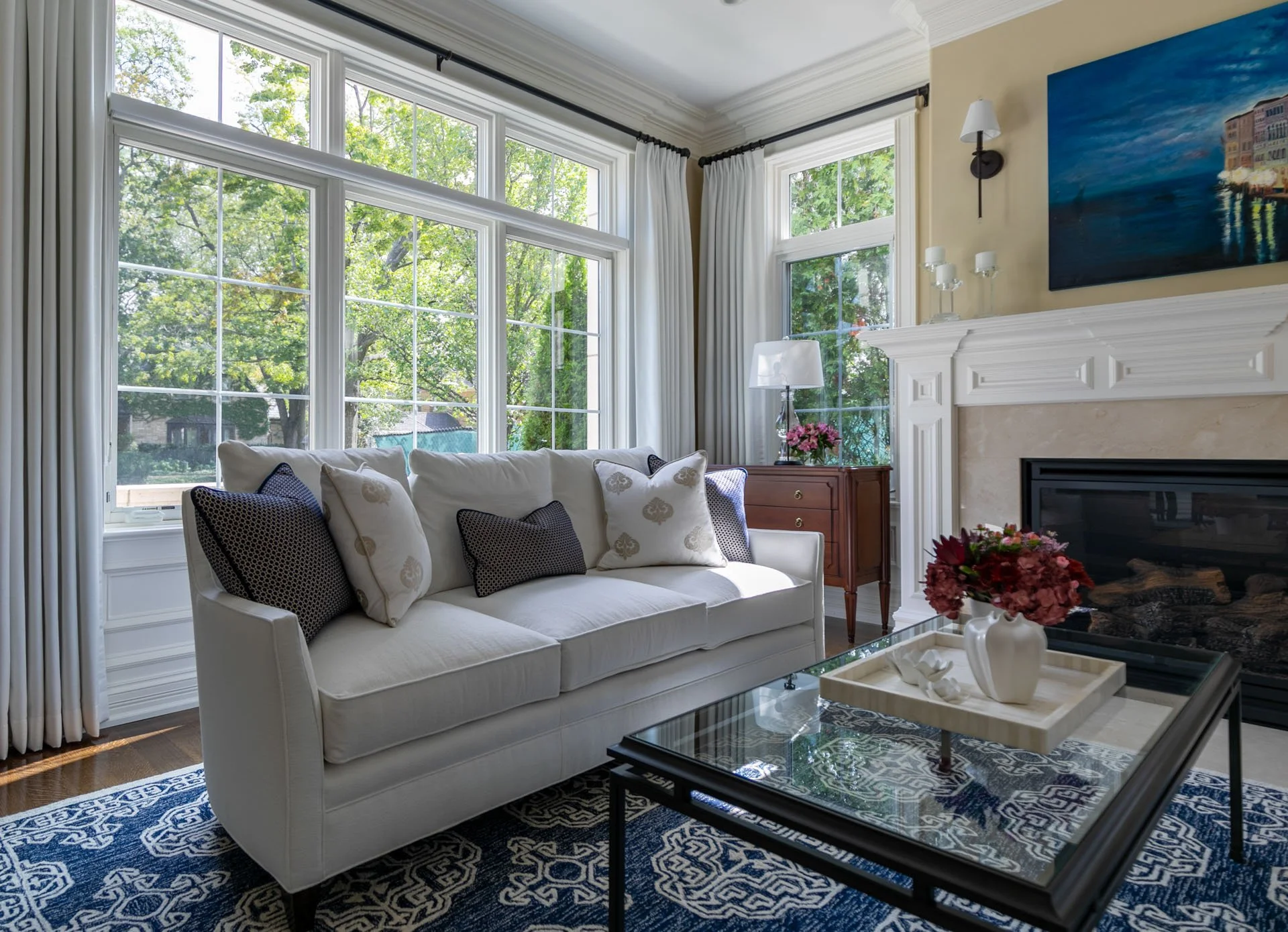 Bright Lincolnwood IL living room with white sofa, navy patterned rug, large windows, and classic fireplace mantel, designed by Wendy Nobriga Interiors.