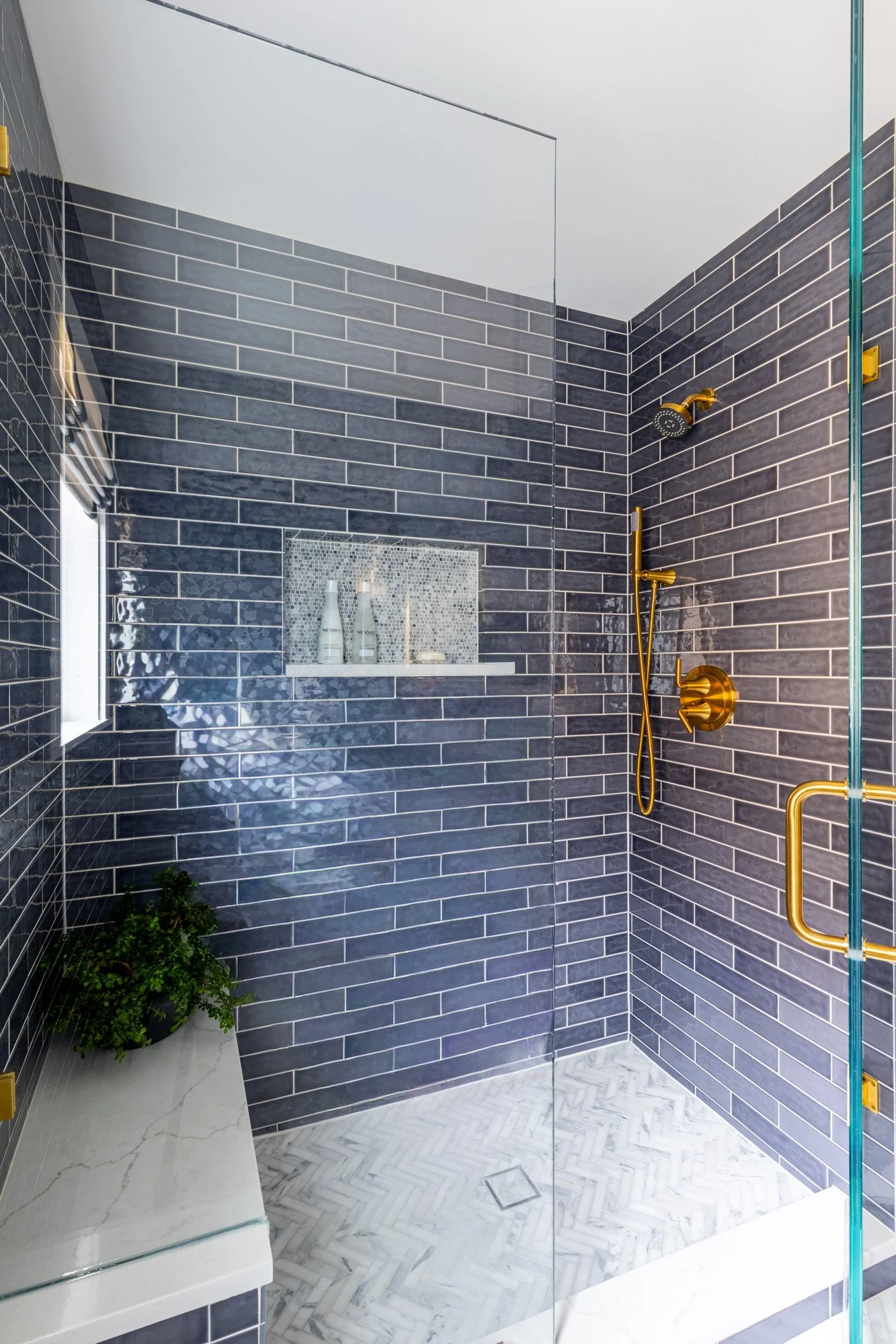 Walk-in shower in a North Edgebrook Chicago first-floor bathroom featuring blue ceramic wall tile, Calacatta Gold marble herringbone mosaic flooring, a frameless glass enclosure, a built-in bench, recessed shower niche, and brass shower fixtures, des
