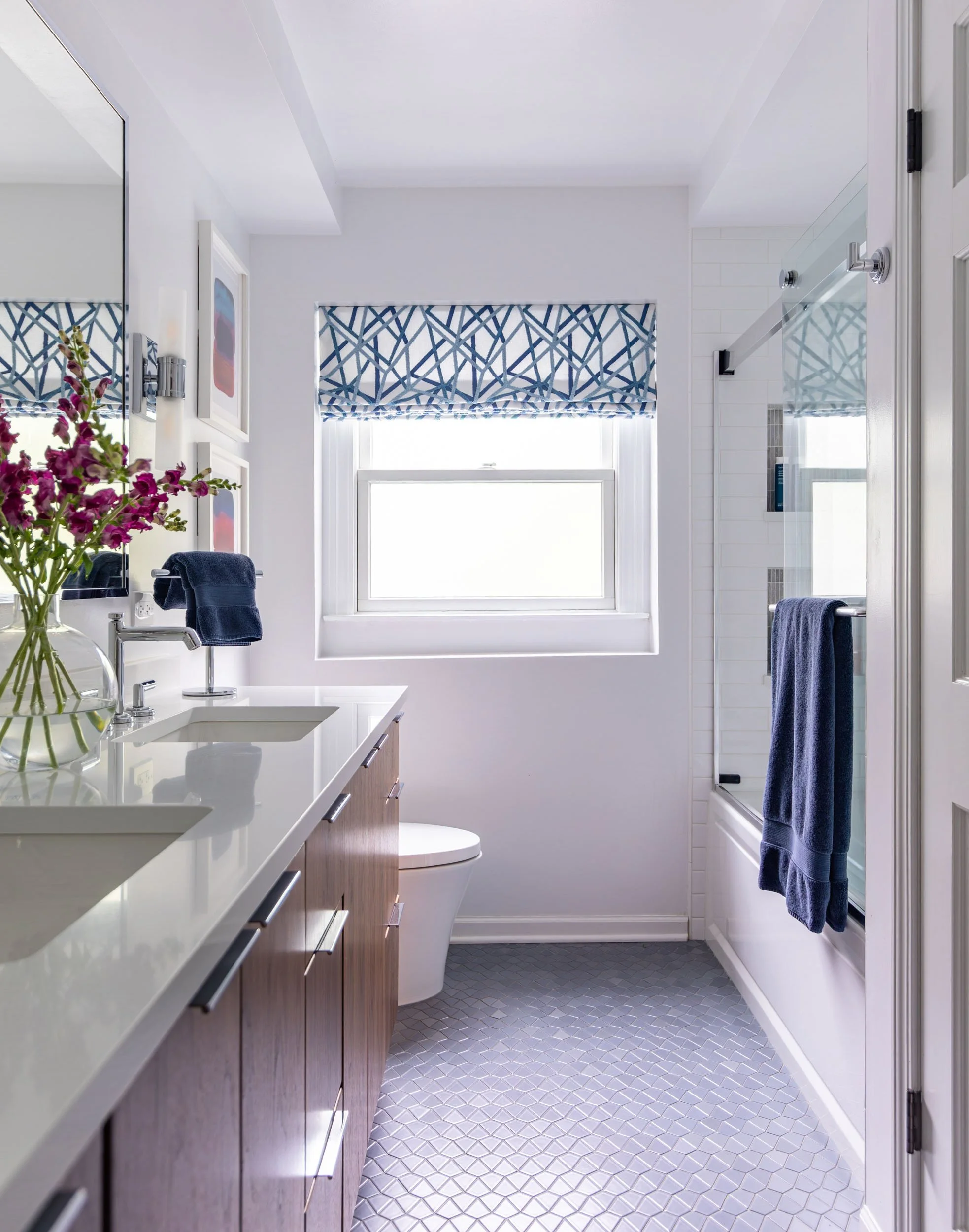 Kids hall bathroom in Northbrook, Illinois with walnut vanity, white quartz countertop, glass tub enclosure, patterned roman shade, and gray mosaic tile floor. Design by Wendy Nobriga Interiors.