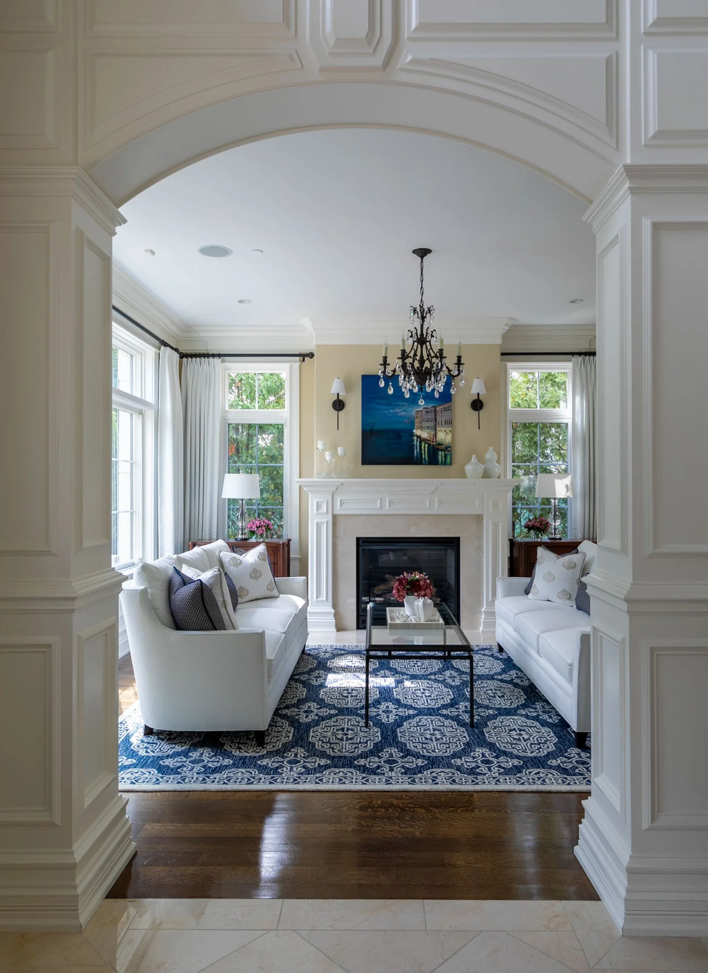 Lincolnwood IL living room view through arched doorway, featuring white sofas, fireplace, chandelier, and blue patterned rug, designed by Wendy Nobriga Interiors.
