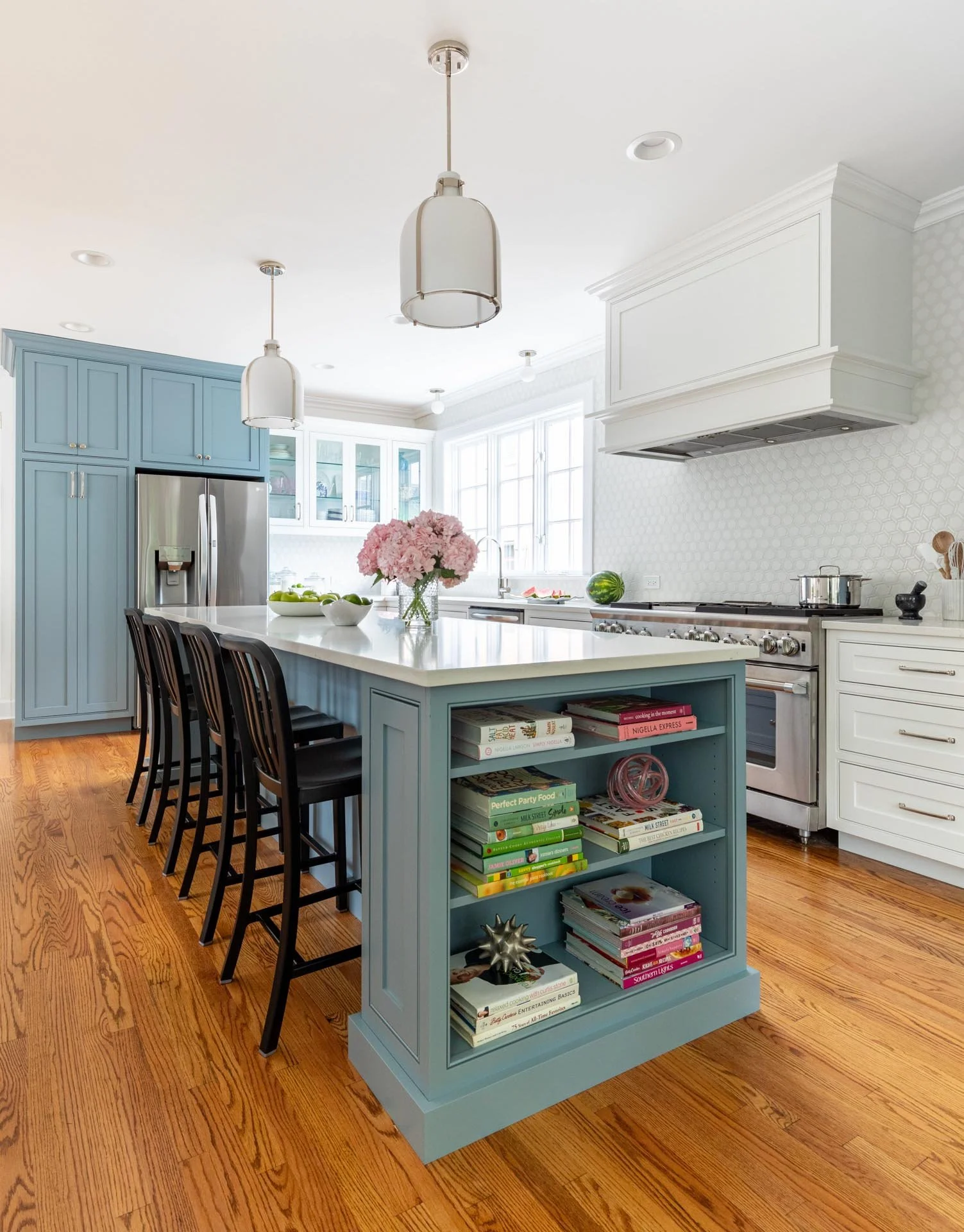 Lake Forest IL transitional kitchen with a white quartz island featuring a built-in bookshelf, blue cabinetry, stainless steel range, and pendant lighting, designed by Wendy Nobriga Interiors.