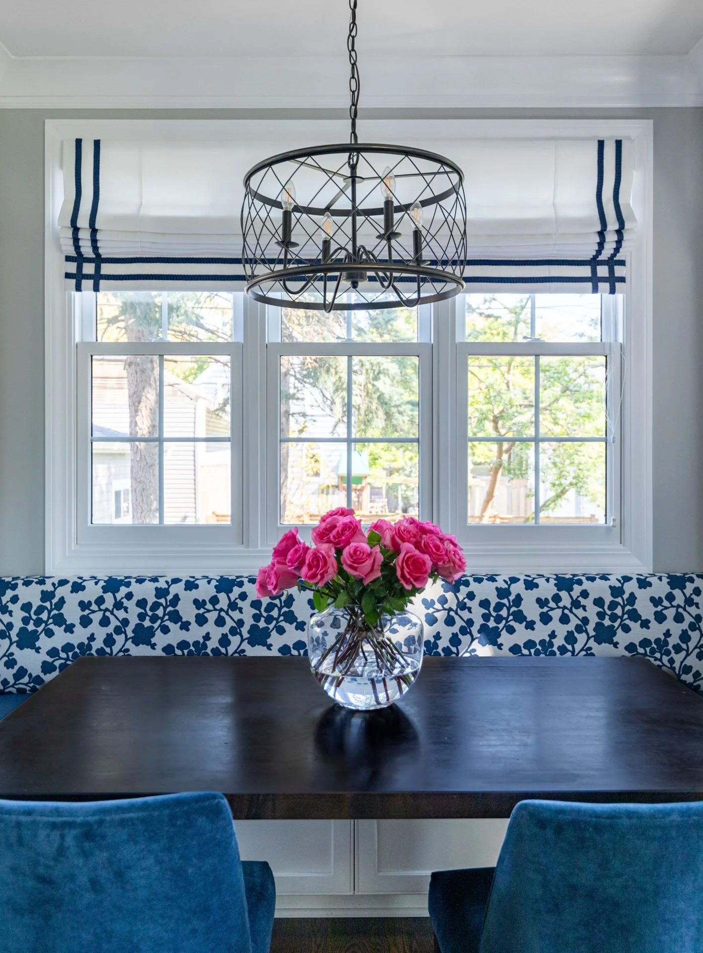 La Grange IL kitchen breakfast nook featuring custom blue floral banquette, Roman shades with navy trim, bronze drum chandelier, and vase of pink roses on wood table by Wendy Nobriga Interiors.