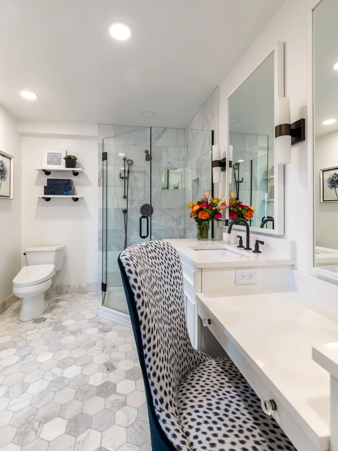 Primary bathroom in Des Plaines IL showcasing a vanity with makeup station, upholstered chair, glass-enclosed shower, and marble hex tile floor, designed by Wendy Nobriga Interiors.