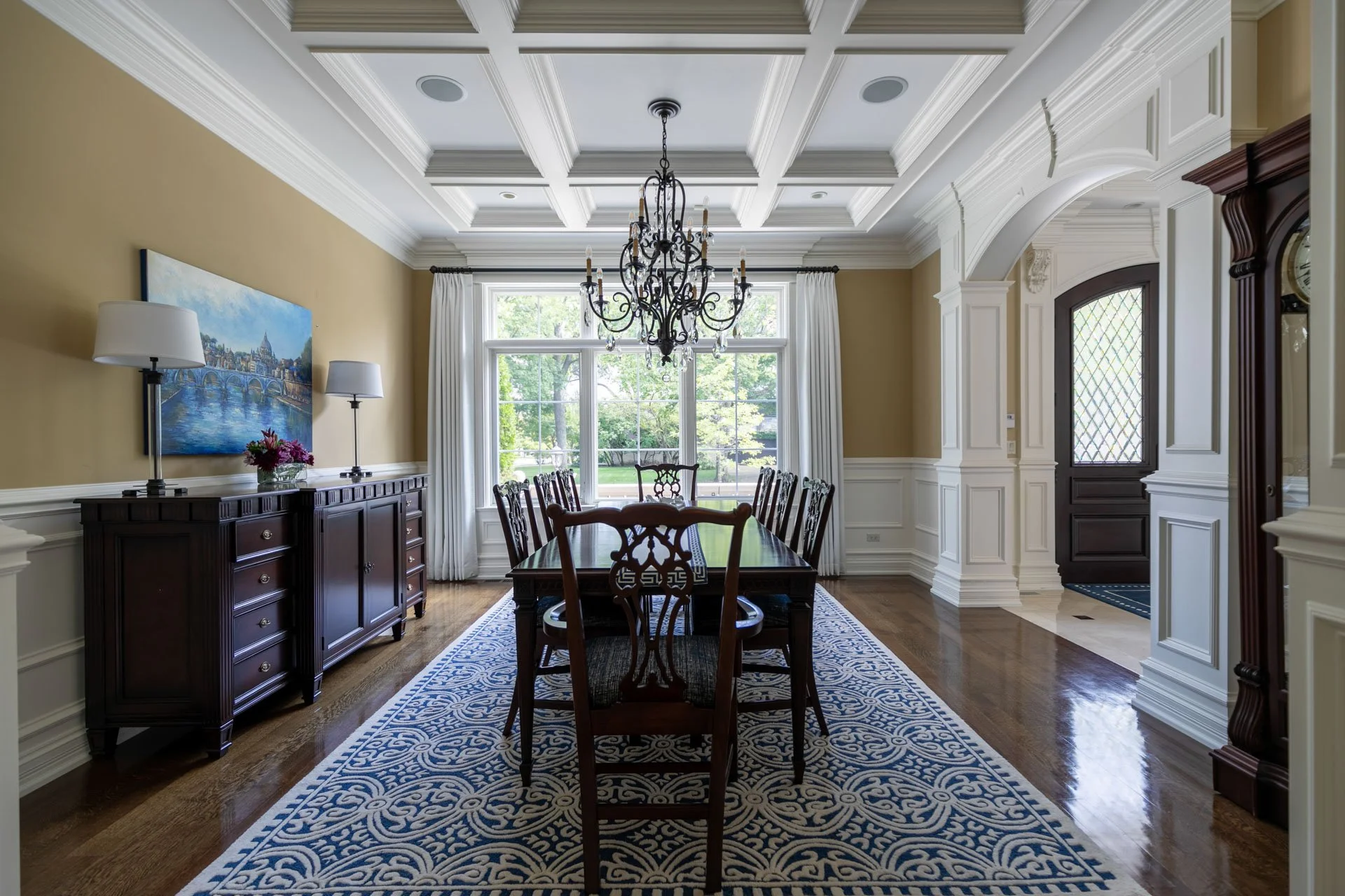 Lincolnwood IL dining room with a coffered ceiling, wrought iron chandelier, long wood dining table, blue patterned rug, and large window overlooking the front yard, designed by Wendy Nobriga Interiors.