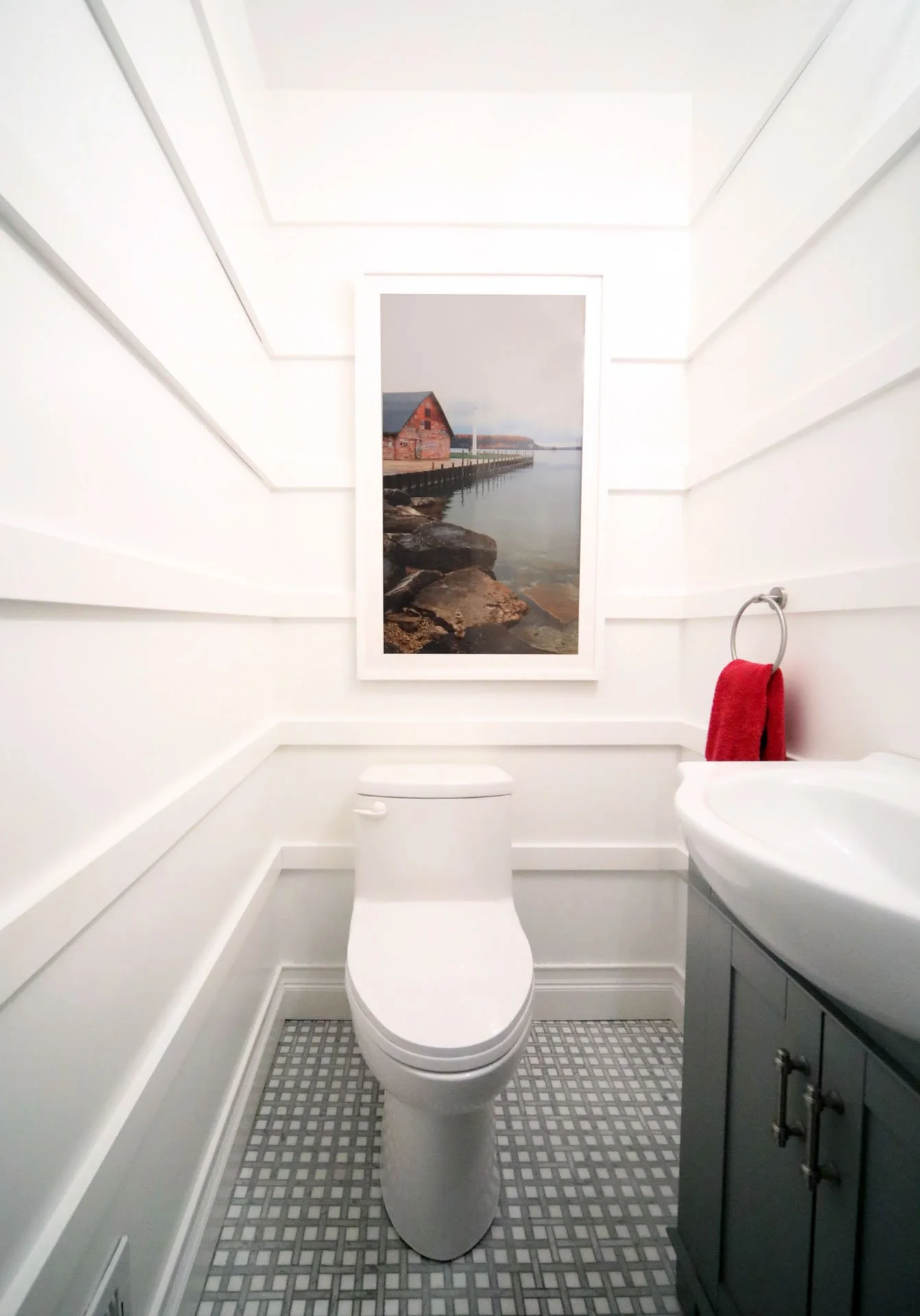 Powder room in Edgebrook, Chicago, IL with white paneled walls, gray vanity, geometric marble tile floor, and wall art above the toilet, designed by Wendy Nobriga Interiors.