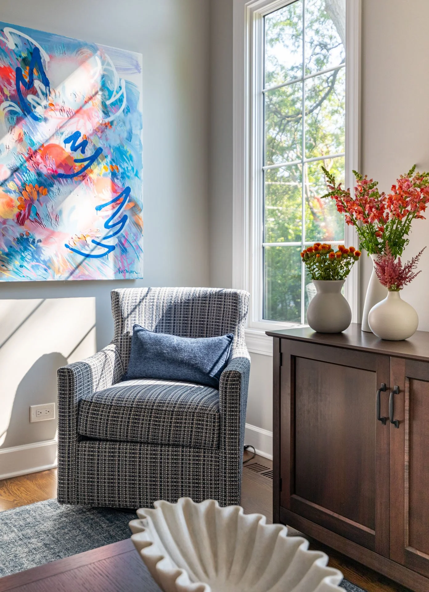 Cozy accent chair in La Grange, IL family room with plaid upholstery, blue pillow, wood console, floral arrangements, and colorful modern artwork, styled by Wendy Nobriga Interiors.