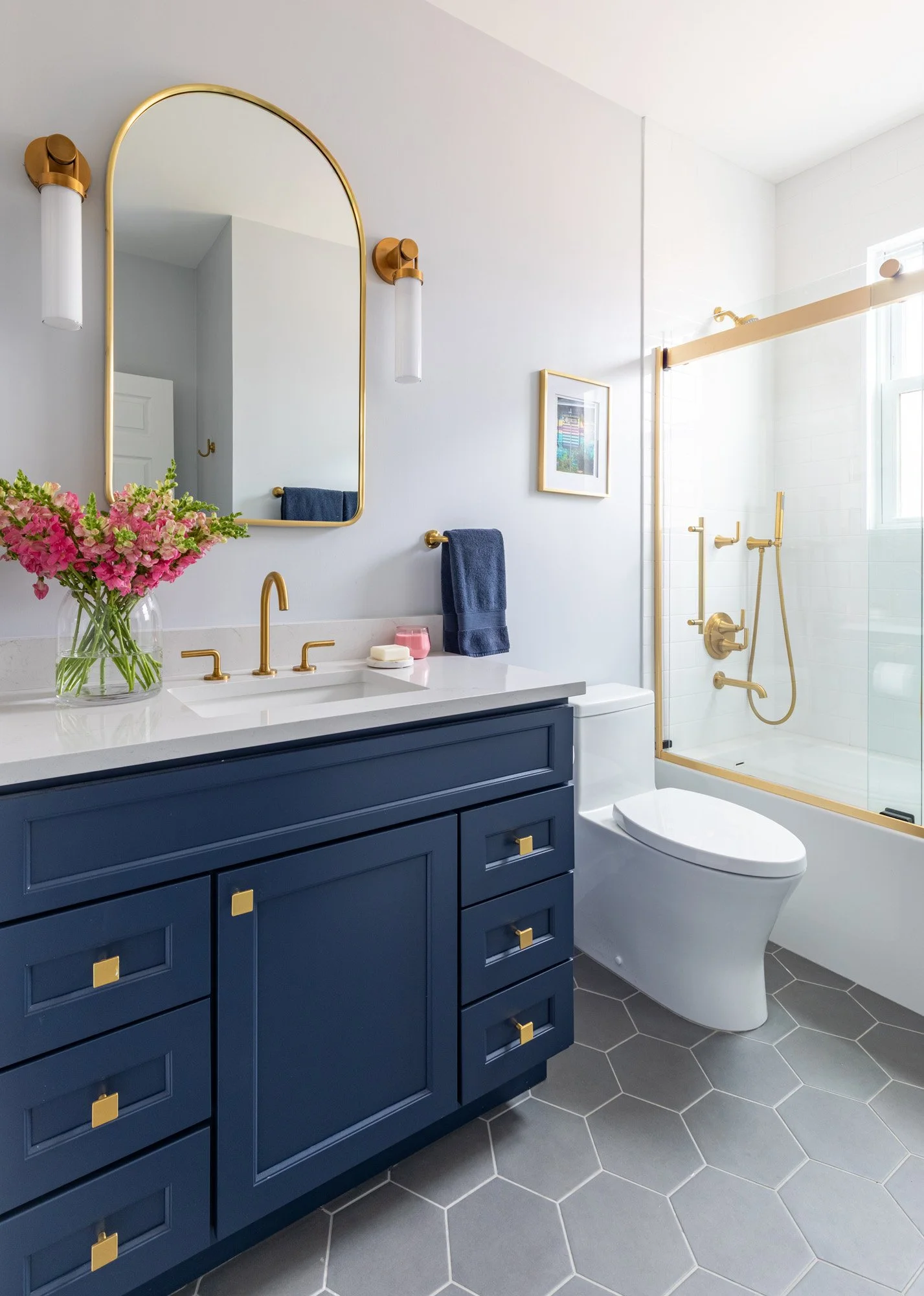 Kids' bathroom in a North Edgebrook Chicago home featuring a custom navy blue vanity, quartz countertop, brass fixtures, hexagon floor tile, and a frameless glass tub shower, designed by Wendy Nobriga Interiors.