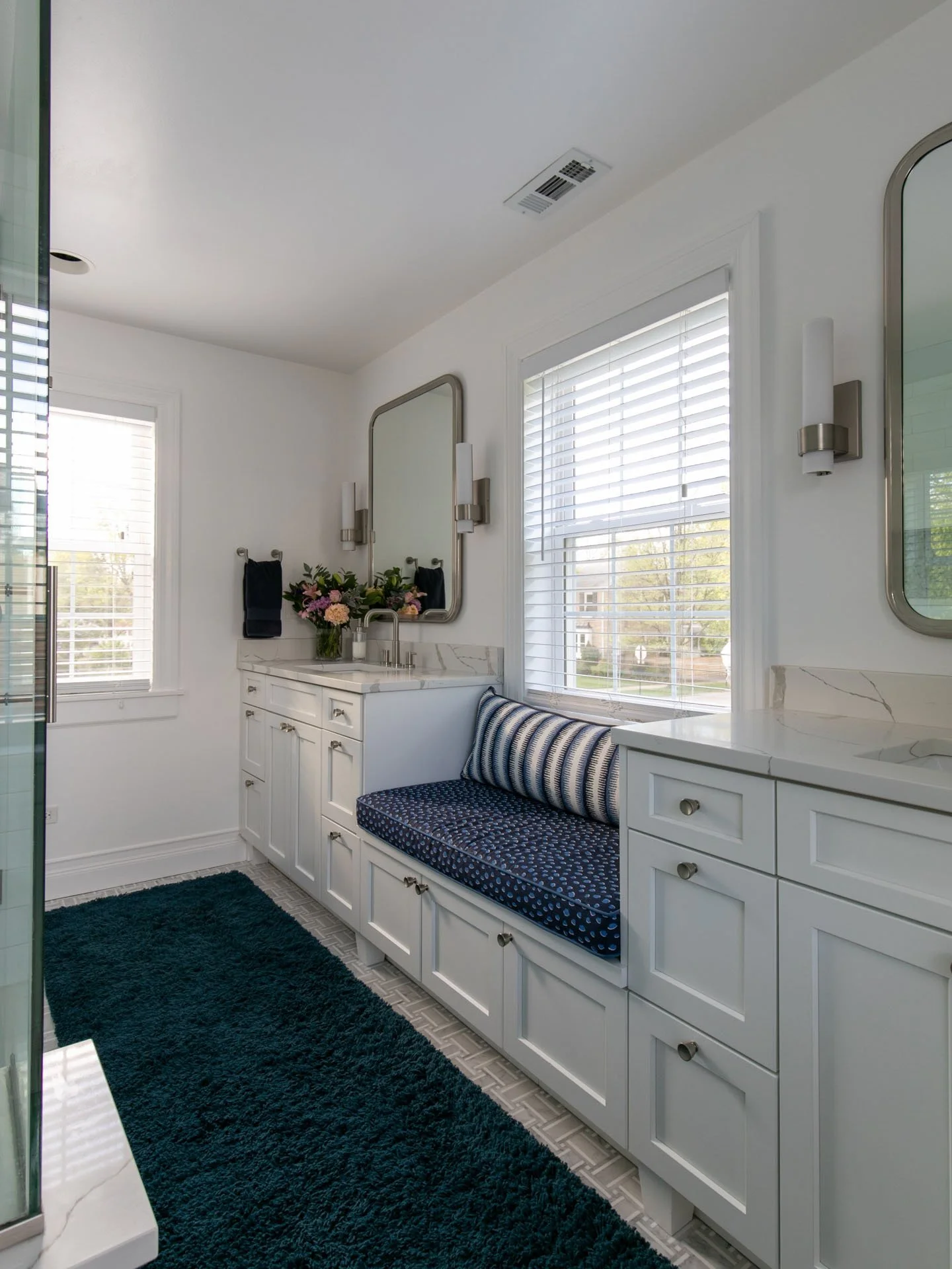 Primary bathroom with double vanity, white quartz countertops, built-in window seat with blue cushion, white cabinetry, and navy rug, Edgebrook, Chicago, IL, designed by Wendy Nobriga Interiors.
