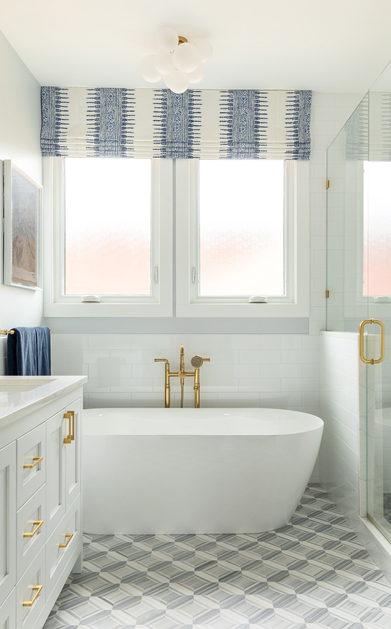 Light-filled primary bathroom in a North Edgebrook Chicago home featuring a freestanding soaking tub, custom white vanity, patterned Roman shades, and marble mosaic flooring, designed by Wendy Nobriga Interiors.