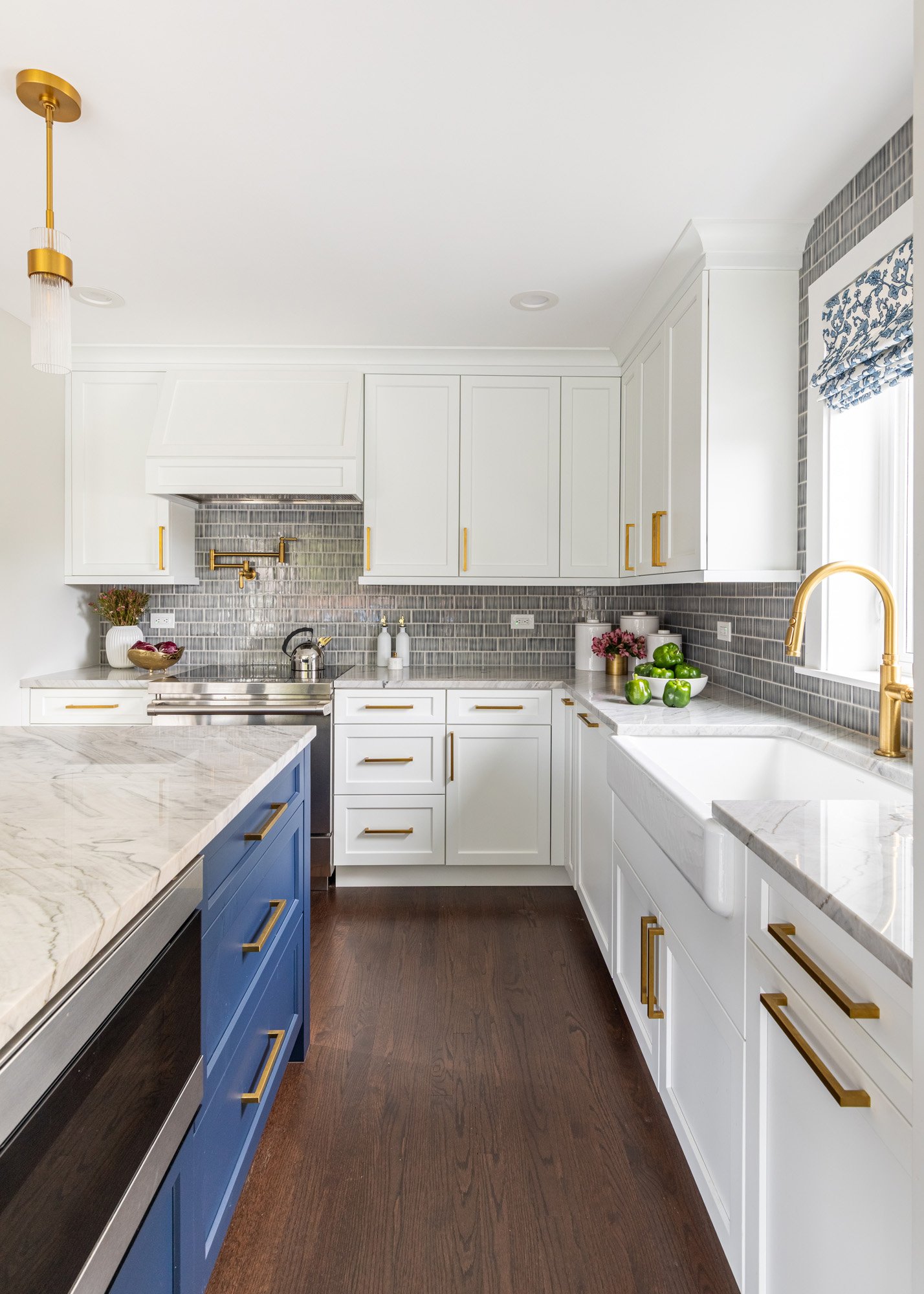 North Edgebrook Chicago kitchen featuring white shaker cabinetry, a large navy blue island, quartzite countertops, a blue handcrafted tile backsplash, farmhouse sink, and brass fixtures, designed by Wendy Nobriga Interiors.