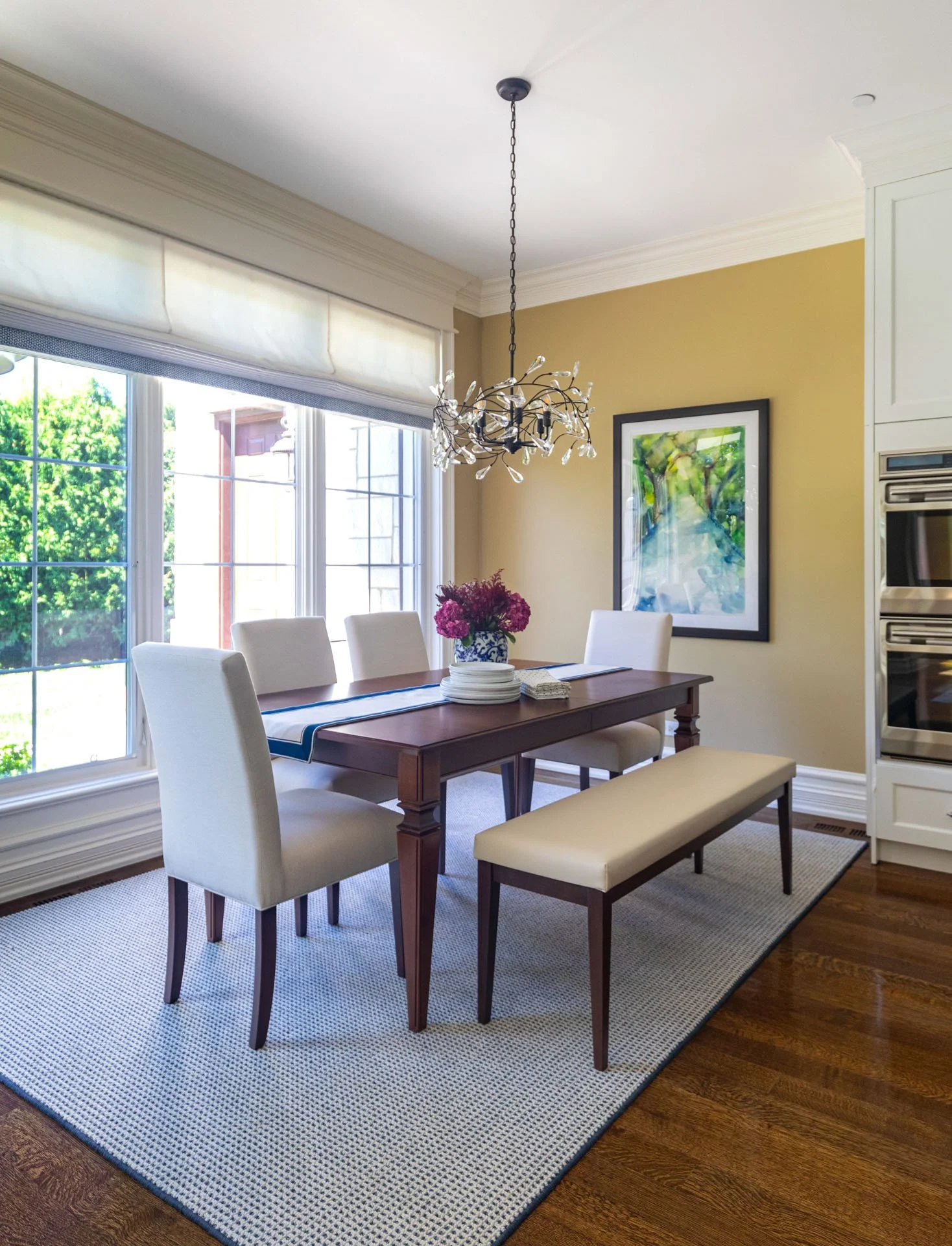 Lincolnwood IL kitchen breakfast nook with a wood dining table, upholstered chairs, a bench, large windows with natural light, and a modern branch-style chandelier, designed by Wendy Nobriga Interiors.