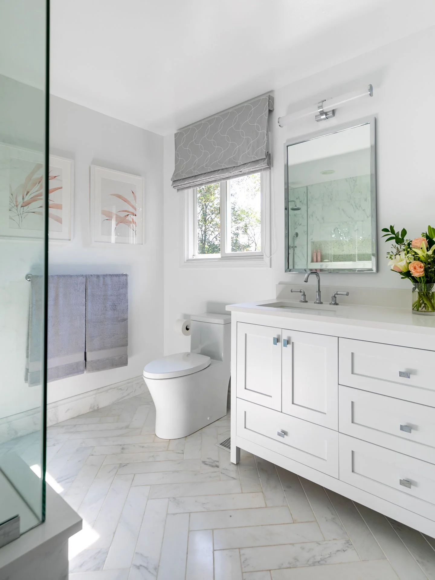 Northbrook IL primary bathroom with white double vanity, modern toilet, herringbone marble floor, gray Roman shade, and wall art above towel bar, designed by Wendy Nobriga Interiors
