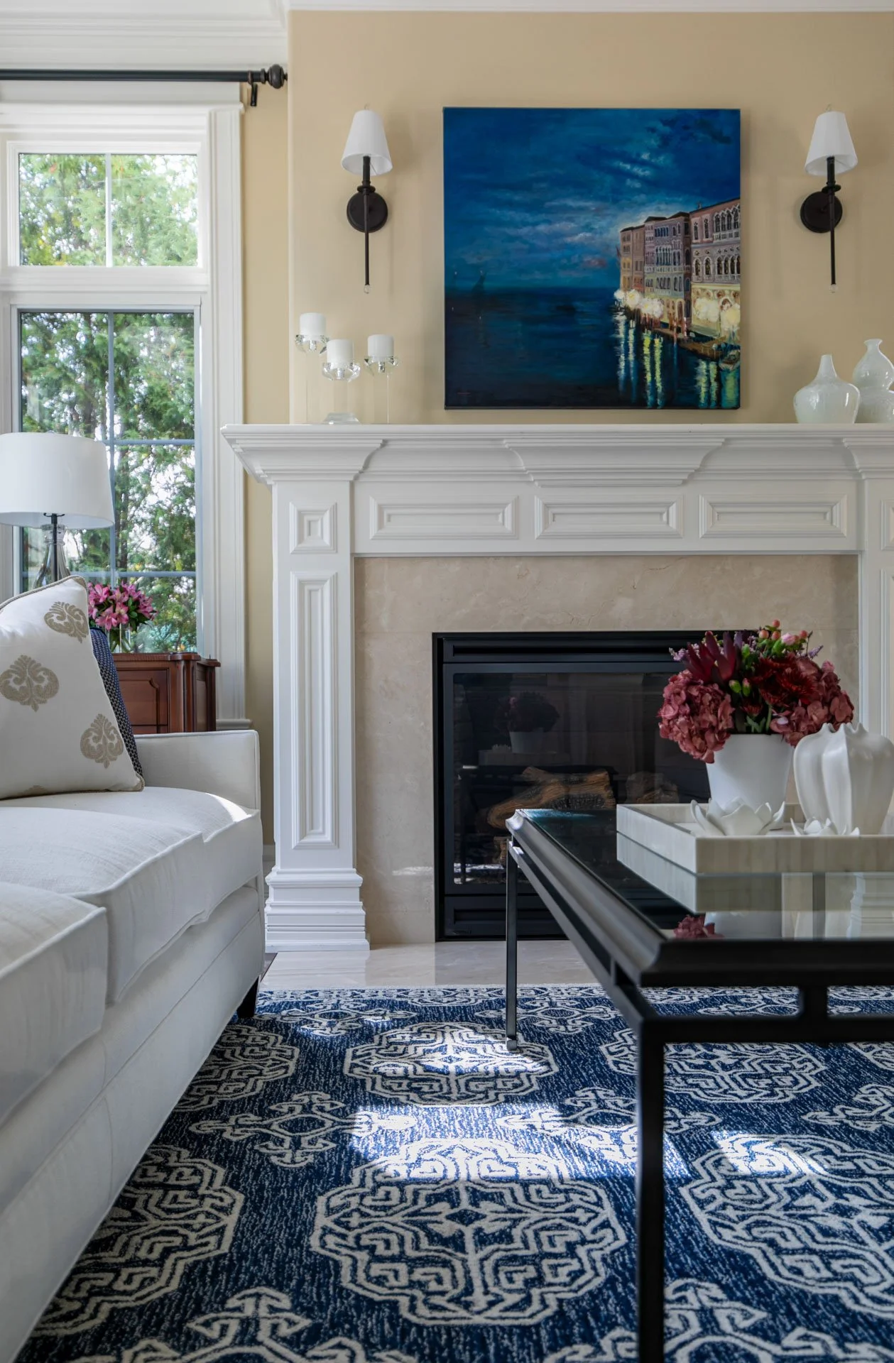 Close-up of Lincolnwood IL living room fireplace with white mantel, modern artwork, navy rug, and floral accents, designed by Wendy Nobriga Interiors.