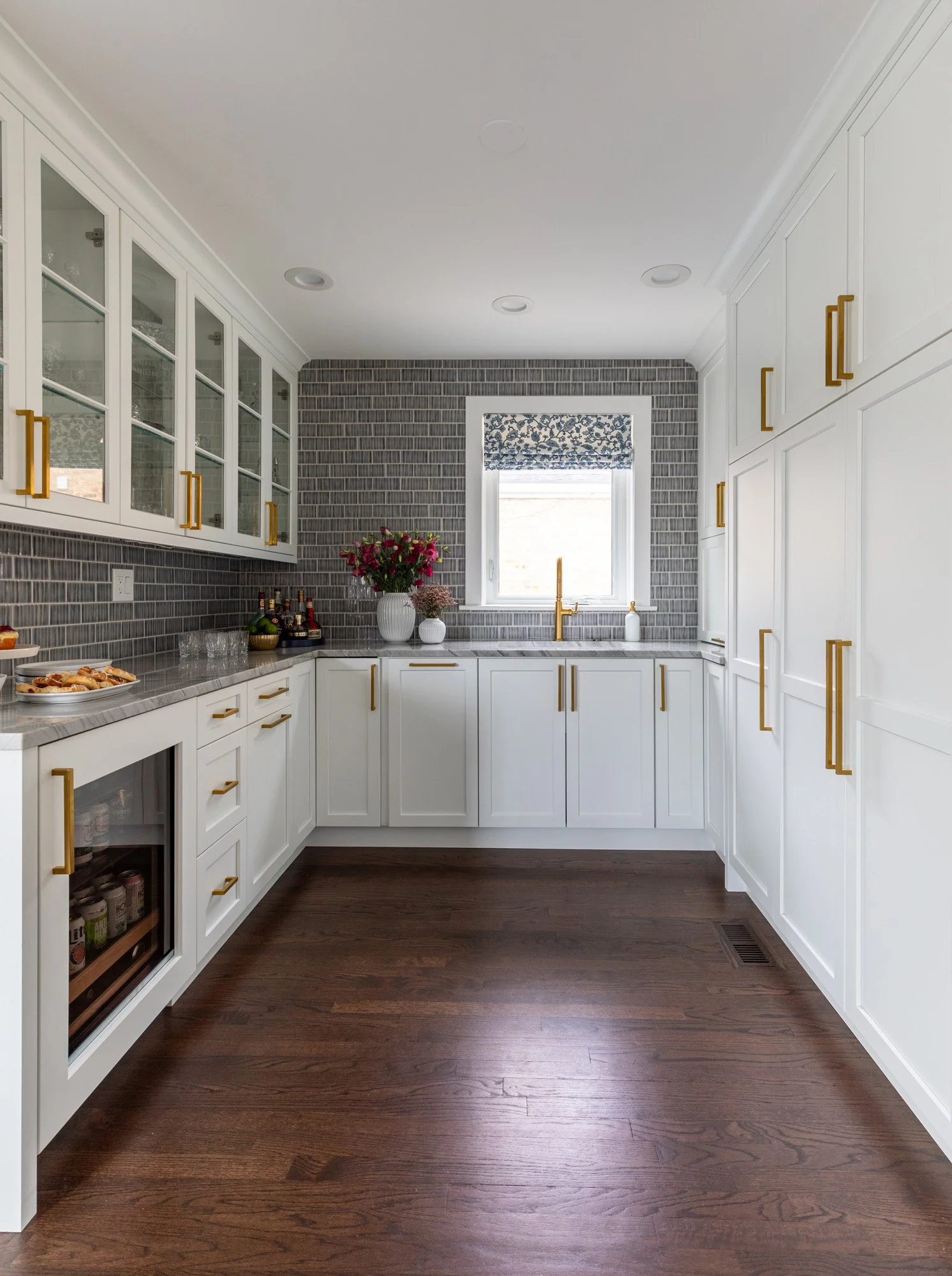 Custom beverage center in a North Edgebrook Chicago home featuring glass display cabinets, a secondary sink, beverage refrigerator, blue handcrafted tile backsplash, quartzite countertops, and custom storage, designed by Wendy Nobriga Interiors.