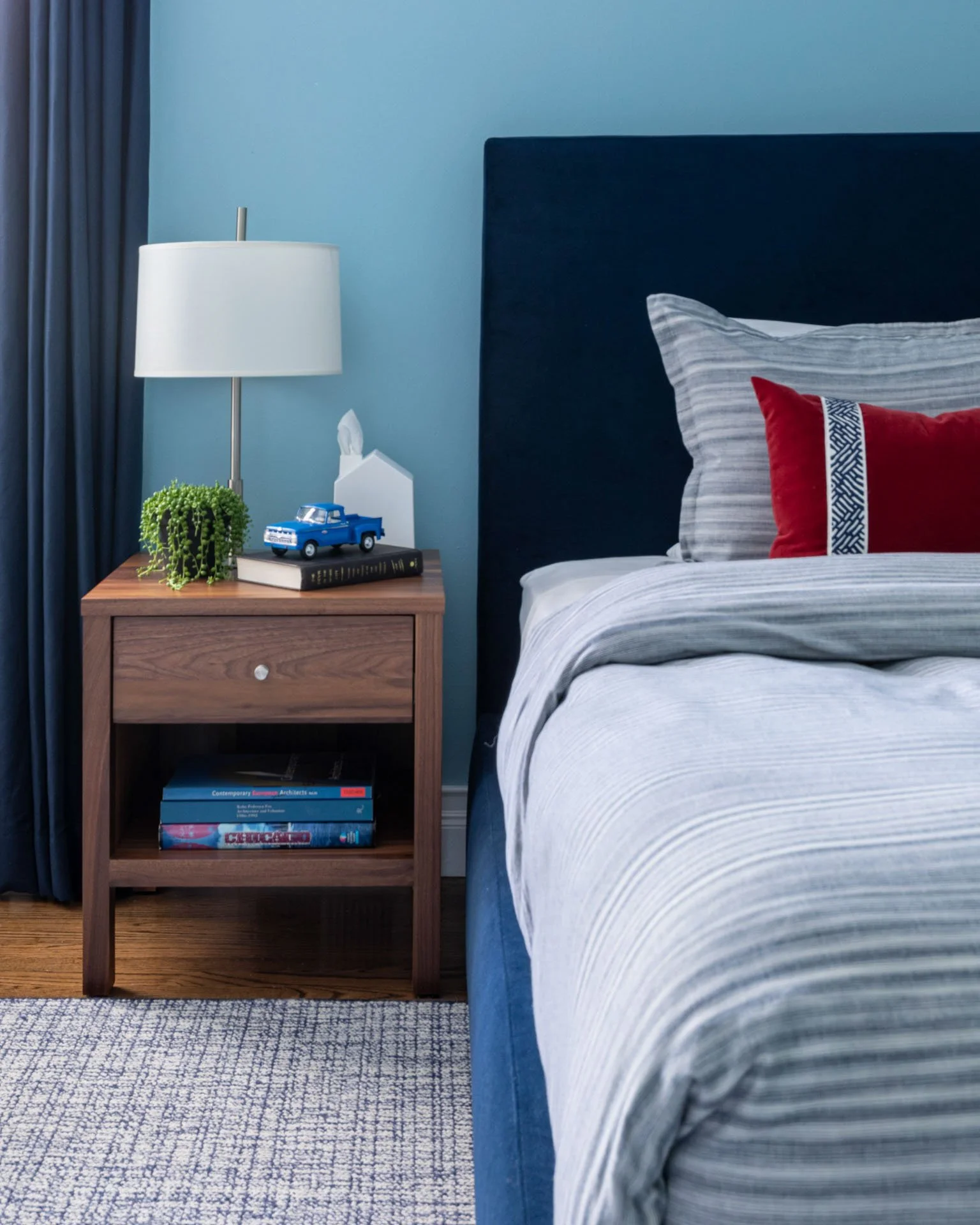 Kid’s bedroom in Edgebrook, Chicago, IL featuring blue upholstered bed with striped bedding, walnut nightstand with lamp and toy truck, designed by Wendy Nobriga Interiors.